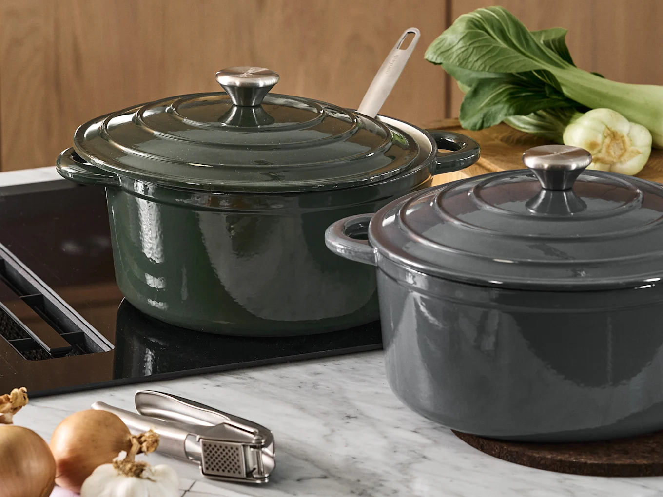 Dos ollas de hierro fundido con tapa, una verde oscuro y otra gris oscuro, en una encimera de cocina con bok choy, cebollas y un prensador de ajos.