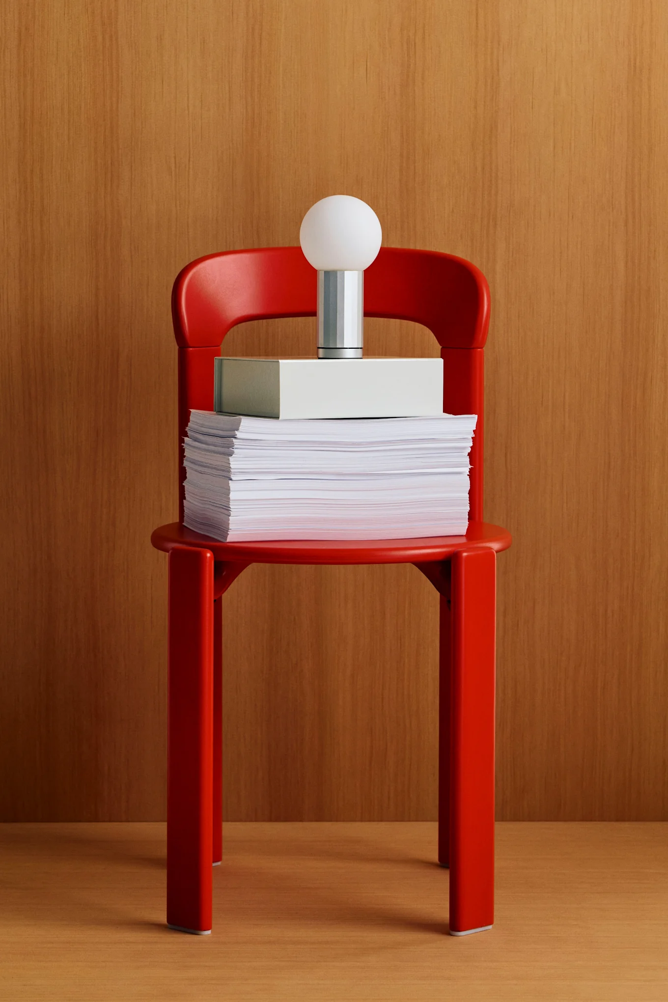Silla roja con una pila de papeles, una caja gris claro y una lámpara moderna con una pantalla esférica blanca sobre un fondo de madera.