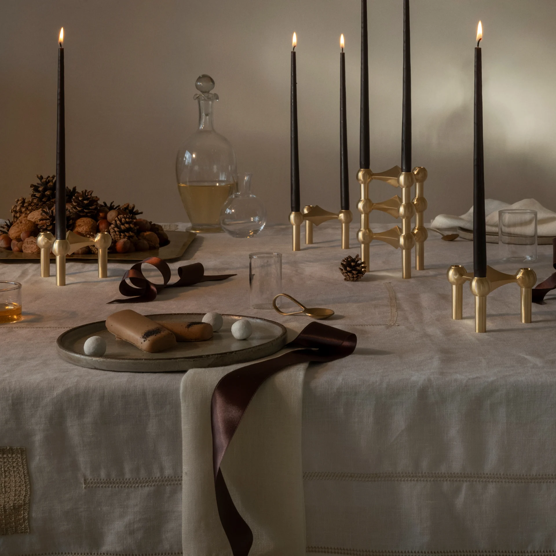 Mesa festiva con velas negras en candelabros modulares de latón, panettone, nueces, piñas y postres.
