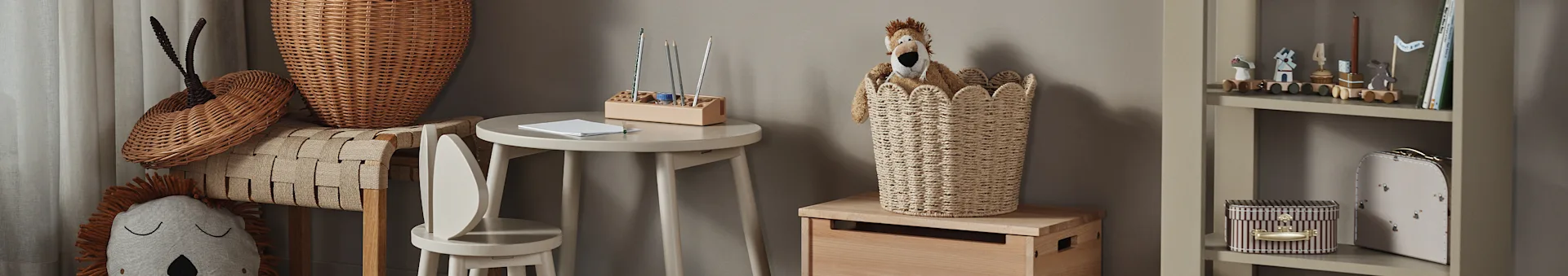 Habitación infantil con un peluche de león en una cesta de mimbre, una pequeña mesa de madera con lápices y una estantería con juguetes y libros.