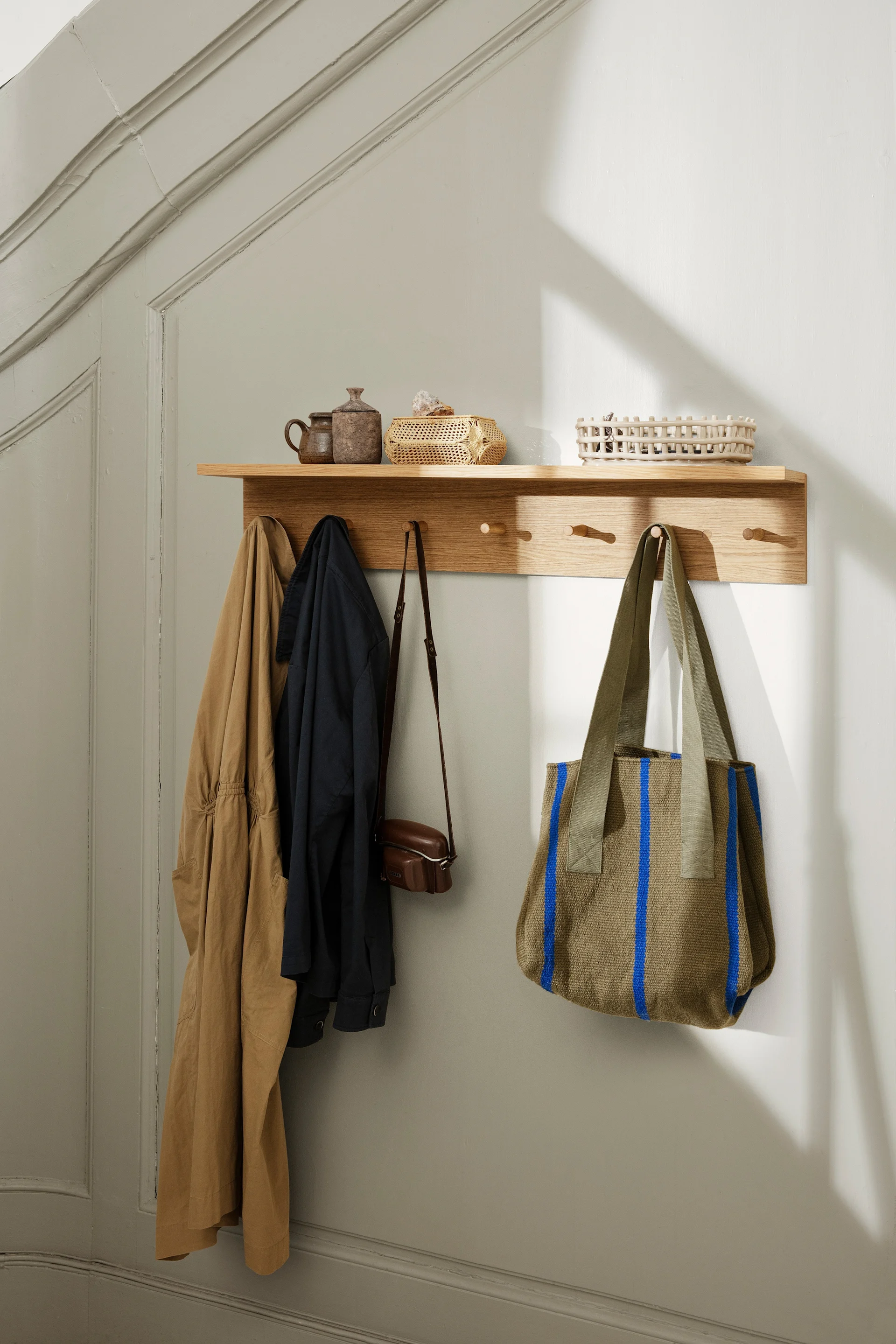 Estante Place Rack con ganchos de madera de Ferm Living. Dos chaquetas, un bolso pequeño de cuero y un bolso de tela verde oliva con rayas azules. En el estante: macetas y cestas. Un estante es perfecto para decorar la pared.