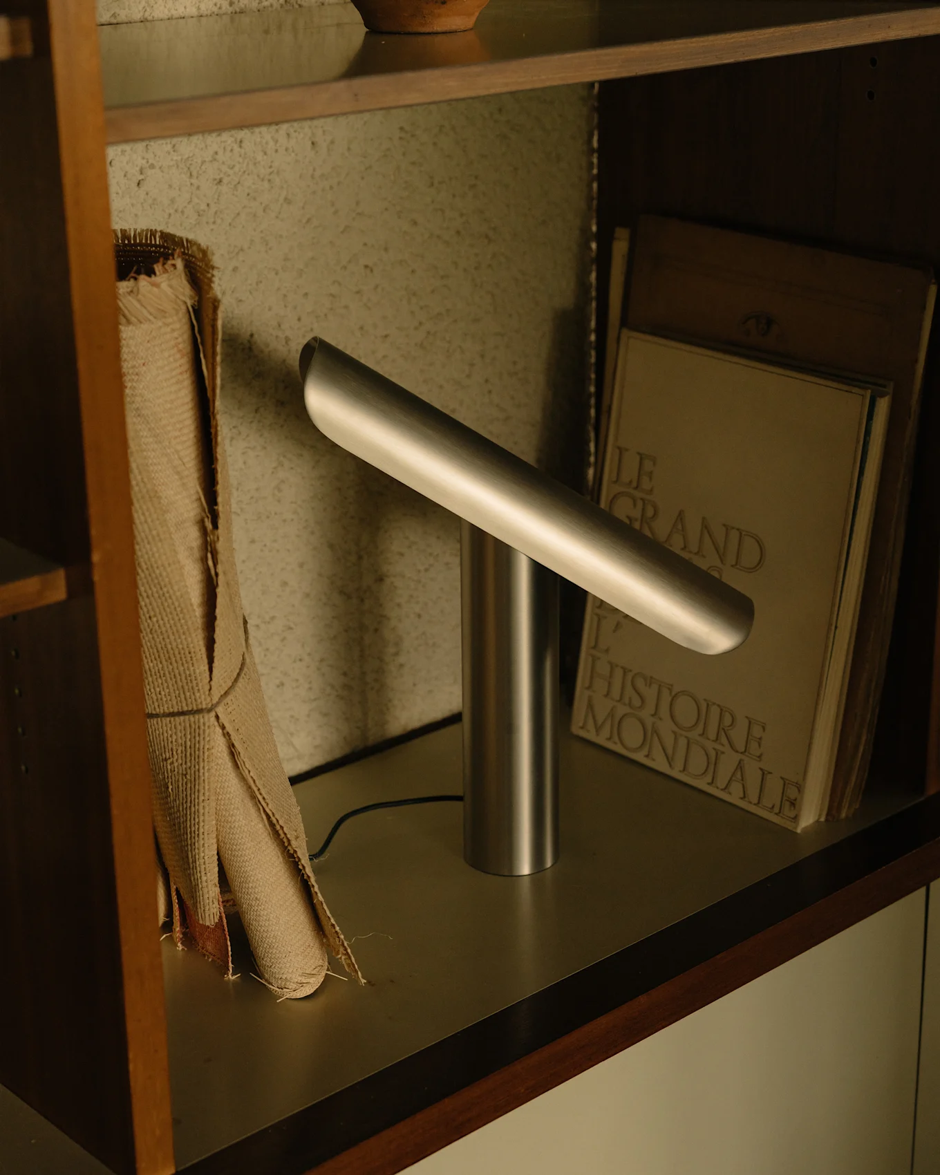 Lámpara de escritorio plateada moderna con base cilíndrica y barra de luz horizontal angulada en un estante.