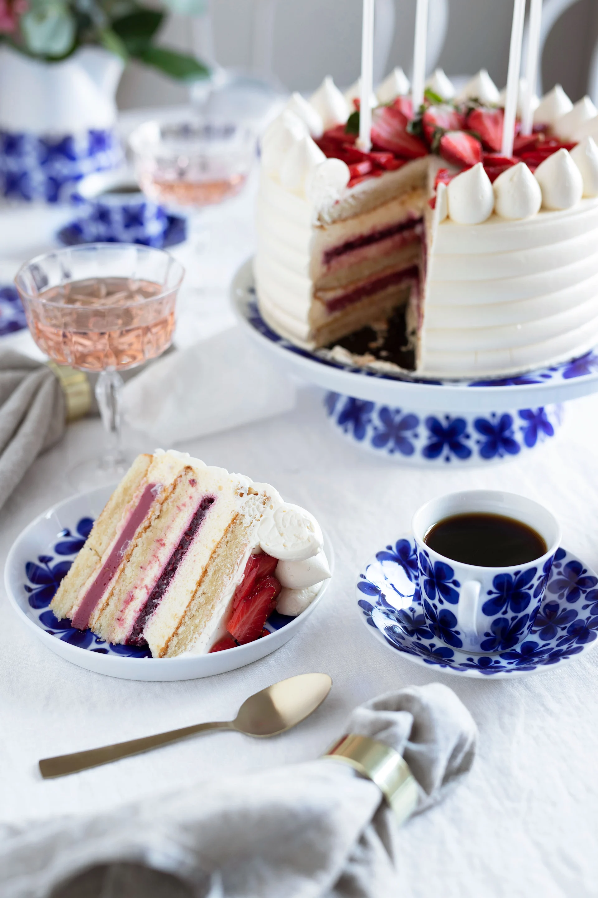 Tarta de fresas con un trozo cortado, servida en vajilla azul y blanca con café y vino rosado.