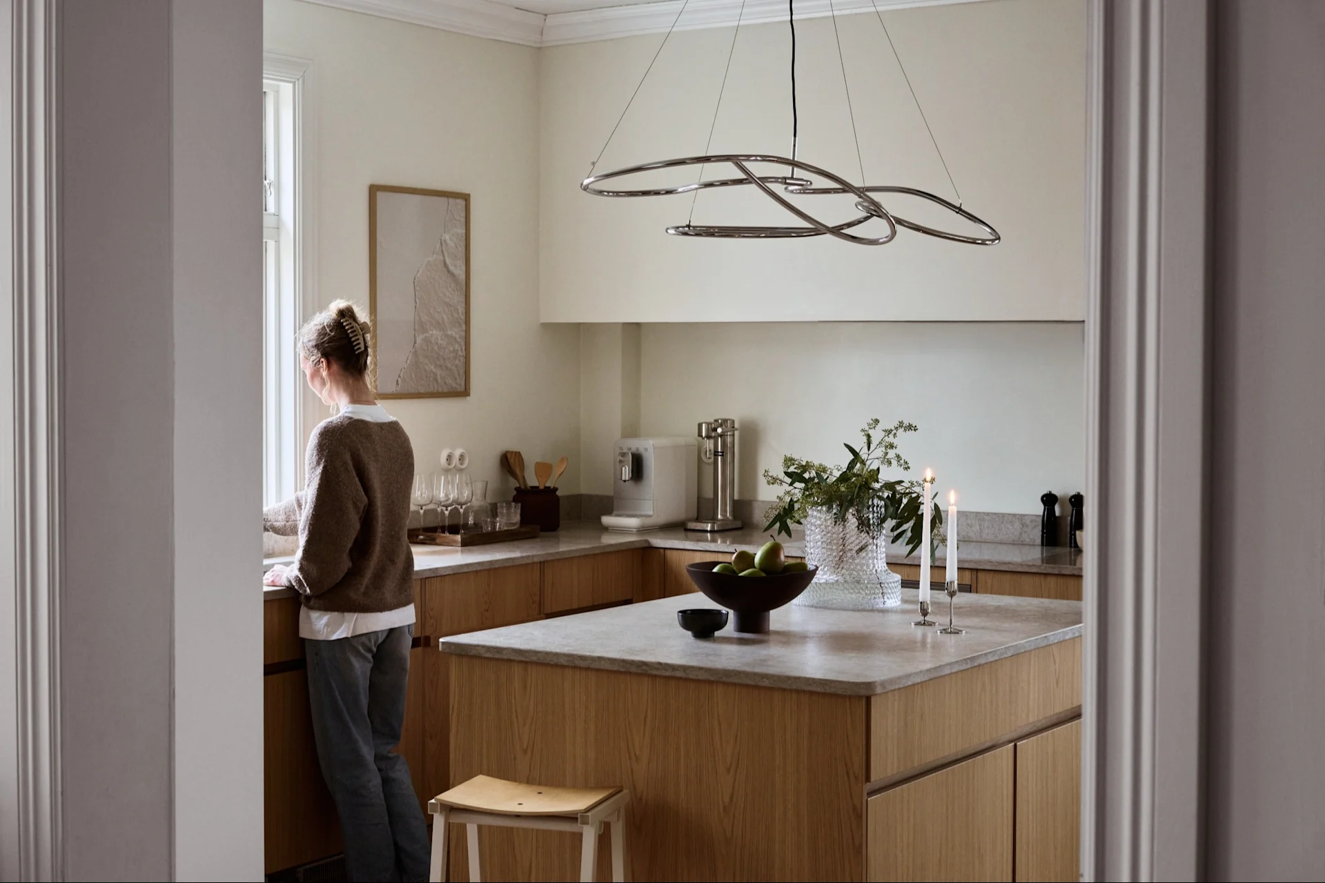 Mujer mirando por la ventana en una cocina moderna y luminosa con armarios de madera clara, encimeras grises y una lámpara colgante escultórica.