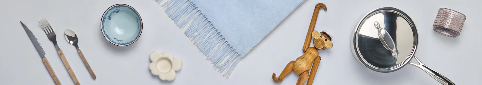 Surtido de artículos para el hogar: cubiertos con mangos de madera, un cuenco azul, posavasos de flores, manta azul, mono de madera, cacerola y vaso.