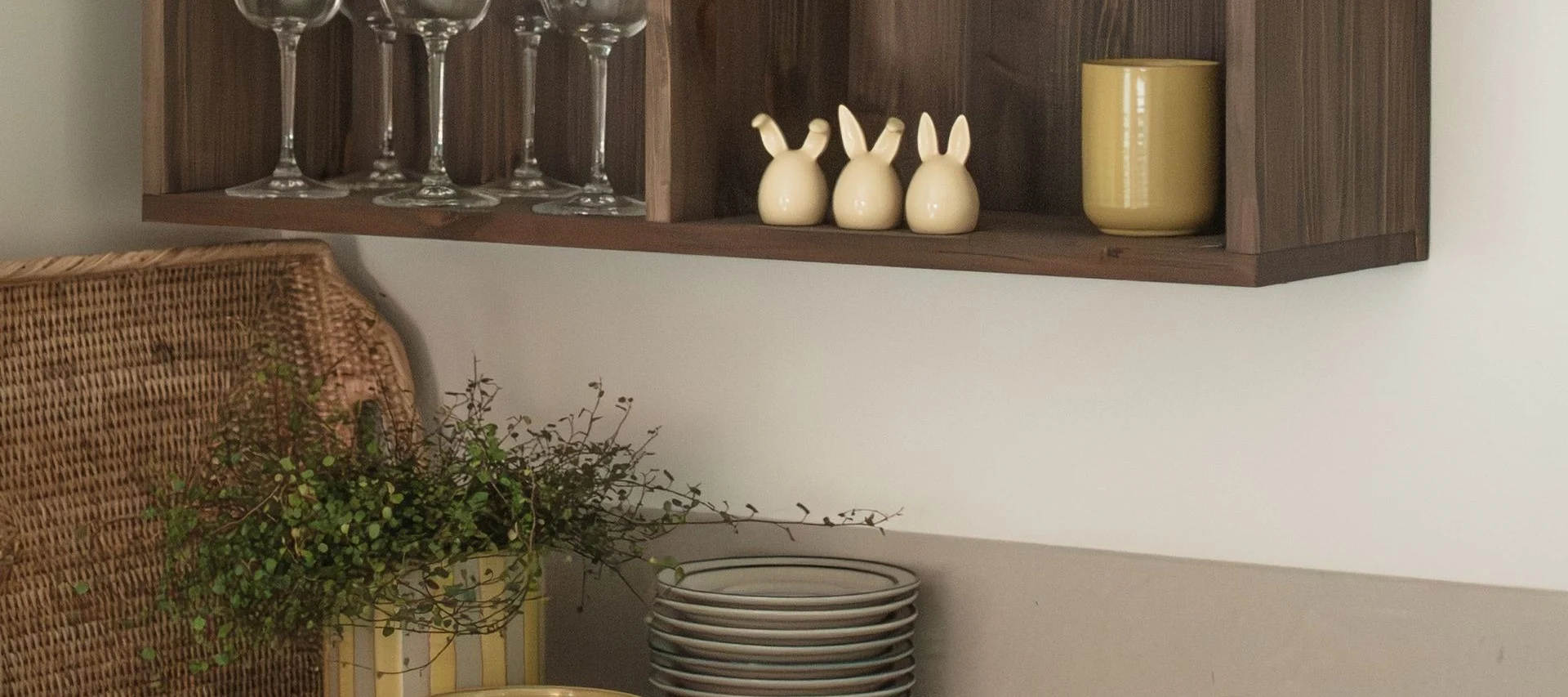 Un estante de madera con copas, tres figuras de conejos y una taza amarilla, con una cesta de mimbre y plantas debajo.