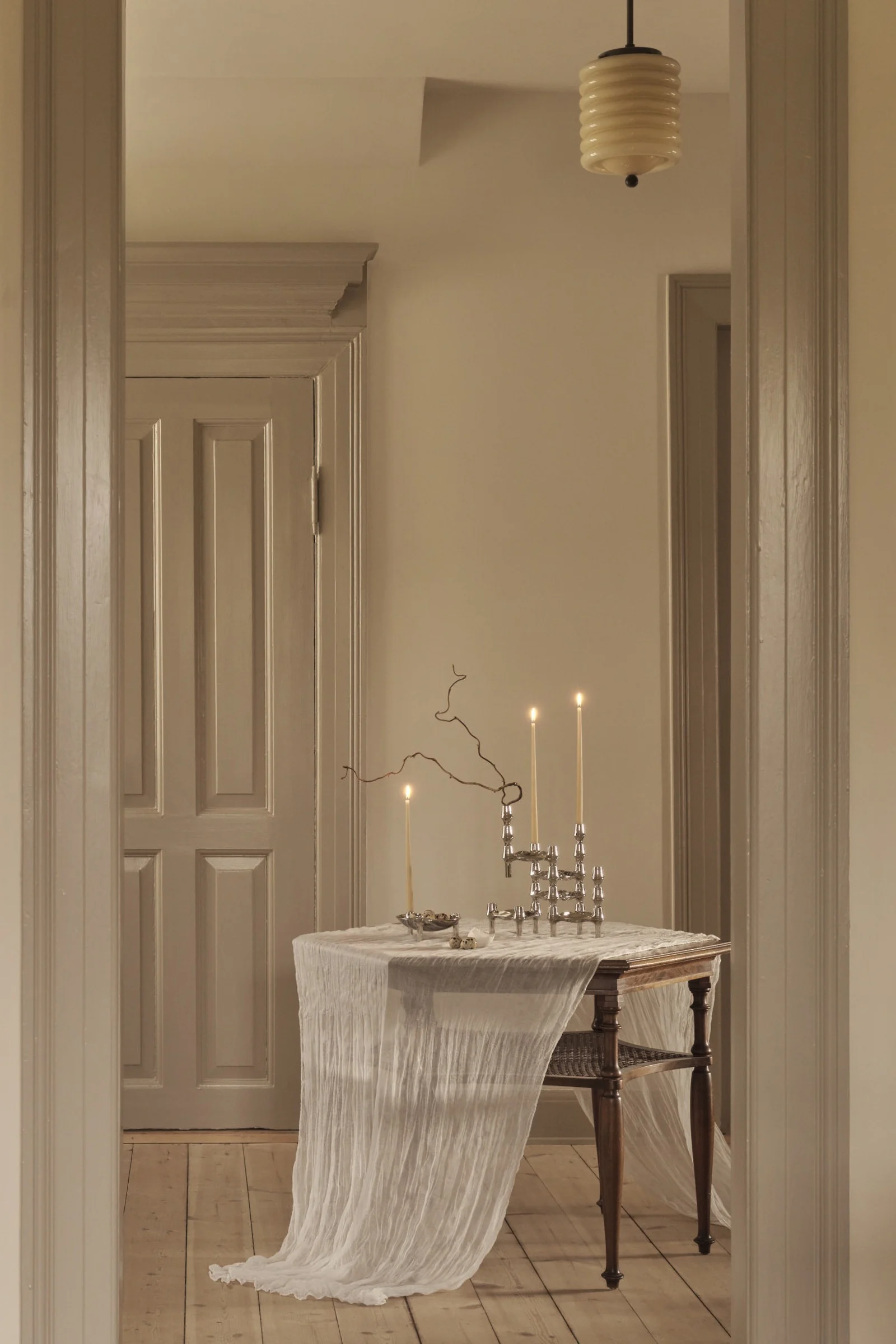 Una mesa de madera con un mantel blanco transparente sostiene tres velas encendidas en candelabros de plata, un cuenco con huevos moteados y una rama en una habitación tenue.