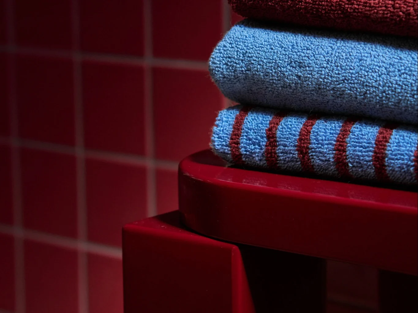 Pila de tres toallas dobladas: rojo oscuro, azul claro y azul con rayas rojas, sobre una superficie roja.