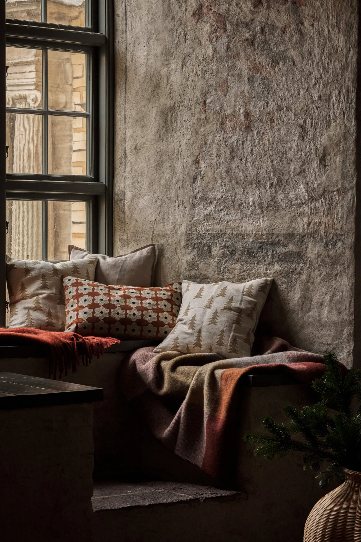 Acogedor asiento junto a la ventana con cojines estampados, mantas cálidas y una rama de pino junto a una pared rústica.