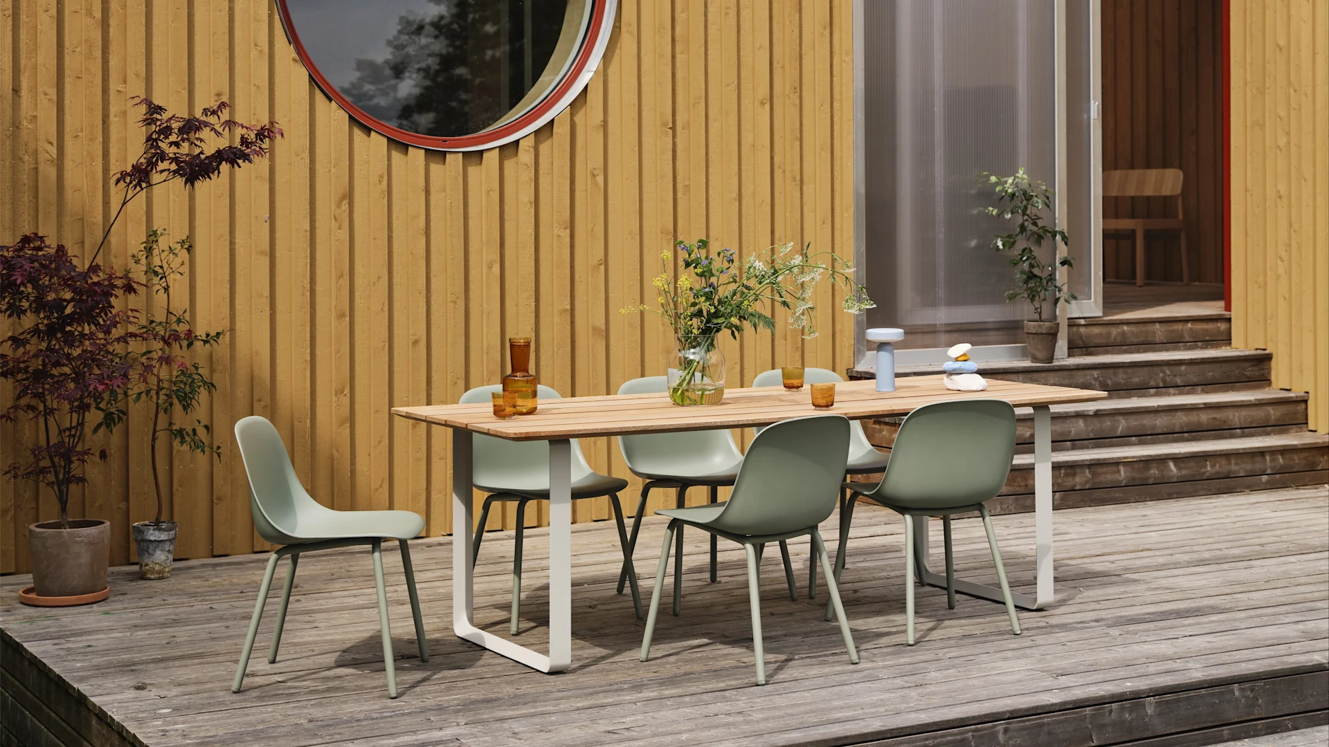 Tendencias en muebles de jardín 2025: las sillas de exterior Fiber verde salvia de Muuto se sitúan en una gran terraza y alrededor de una mesa, con una casa de madera amarilla al fondo.