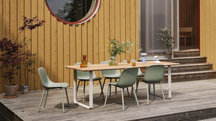 Tendencias en muebles de jardín 2025: las sillas de exterior Fiber verde salvia de Muuto se sitúan en una gran terraza y alrededor de una mesa, con una casa de madera amarilla al fondo.