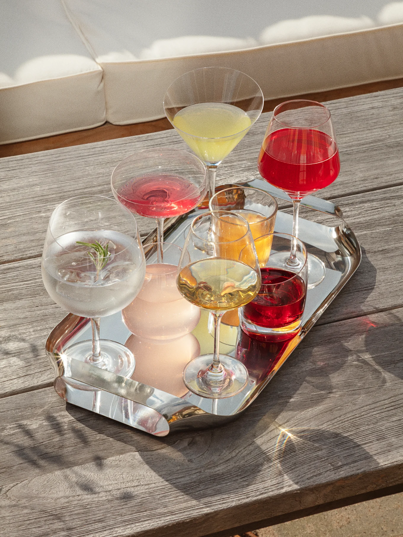 Una bandeja plateada con varias bebidas coloridas sobre una mesa de madera, con un sofá blanco al fondo, bañada por la luz del sol.