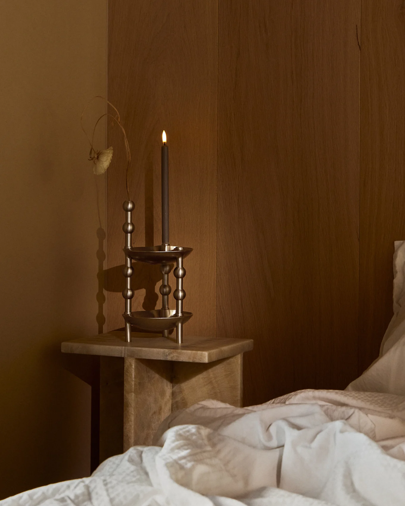Una vela oscura encendida en un portavelas metálico ornamentado sobre una mesita de noche de madera junto a una cama con ropa de cama blanca en una habitación tenue.