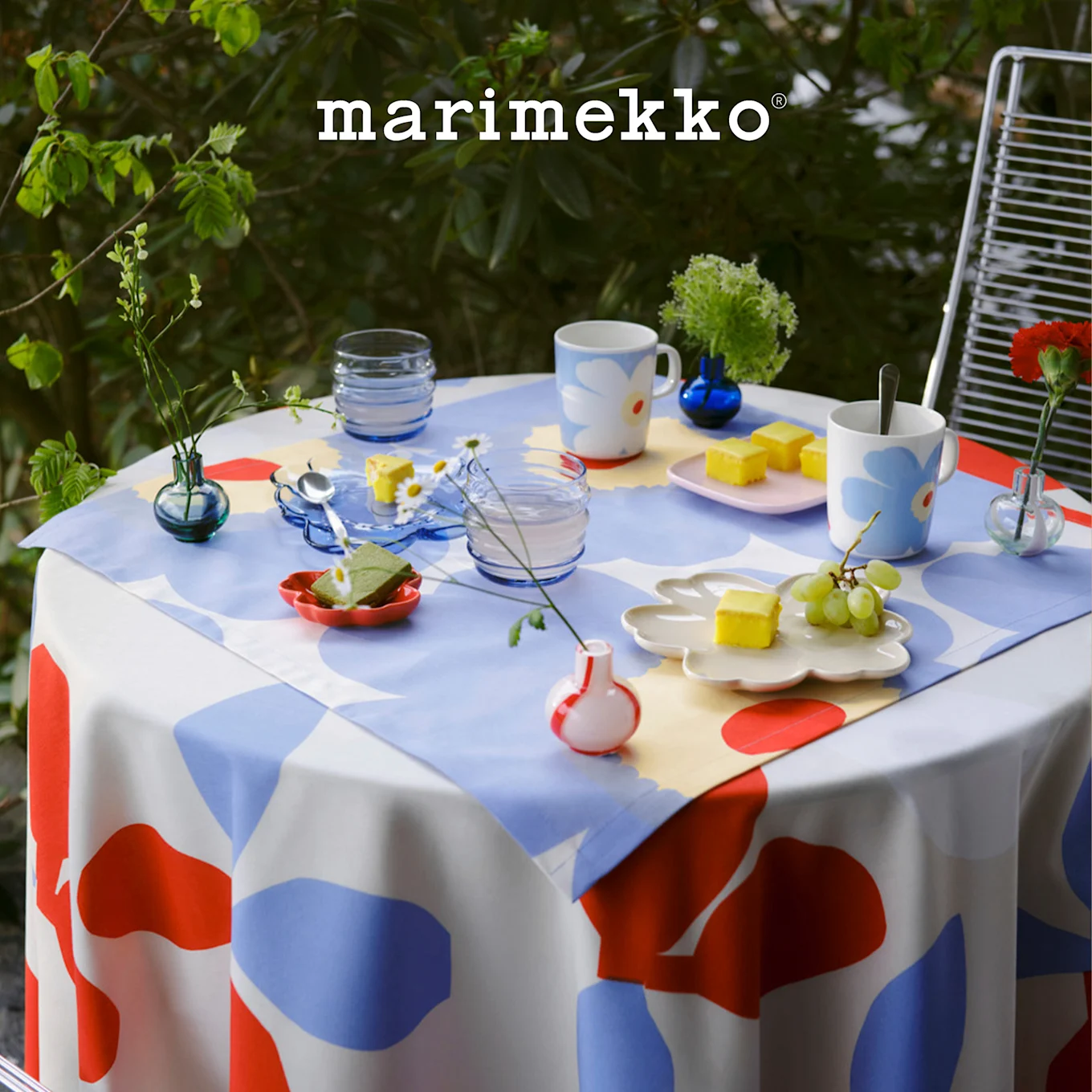 Mesa de exterior con mantel Marimekko estampado en rojo, azul, blanco, con tazas florales, postres y plantas pequeñas. Logotipo de Marimekko.