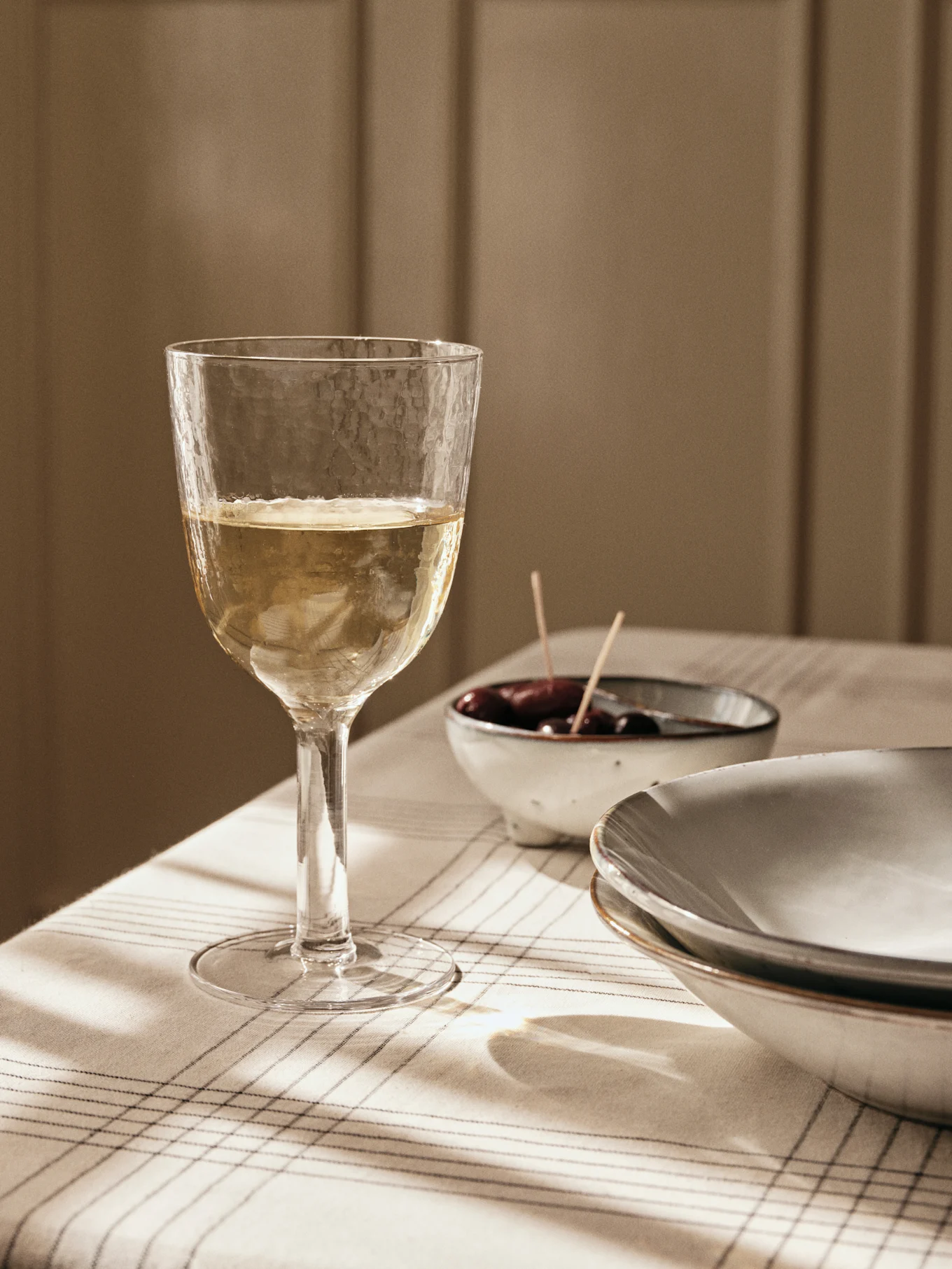 Una copa de vino blanco texturizada sobre un mantel a rayas, con aceitunas y platos al fondo, bañada por la luz de la ventana.