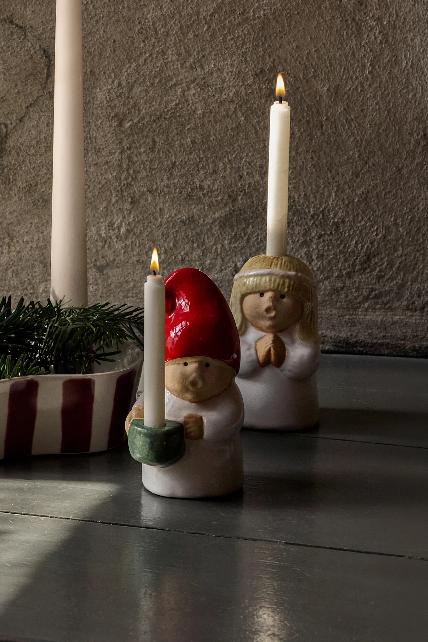 Arreglo de Adviento: cuatro velas sin encender en un portavelas a rayas con pino, flanqueadas por figuras de cerámica (ángel y gnomo) con velas encendidas.