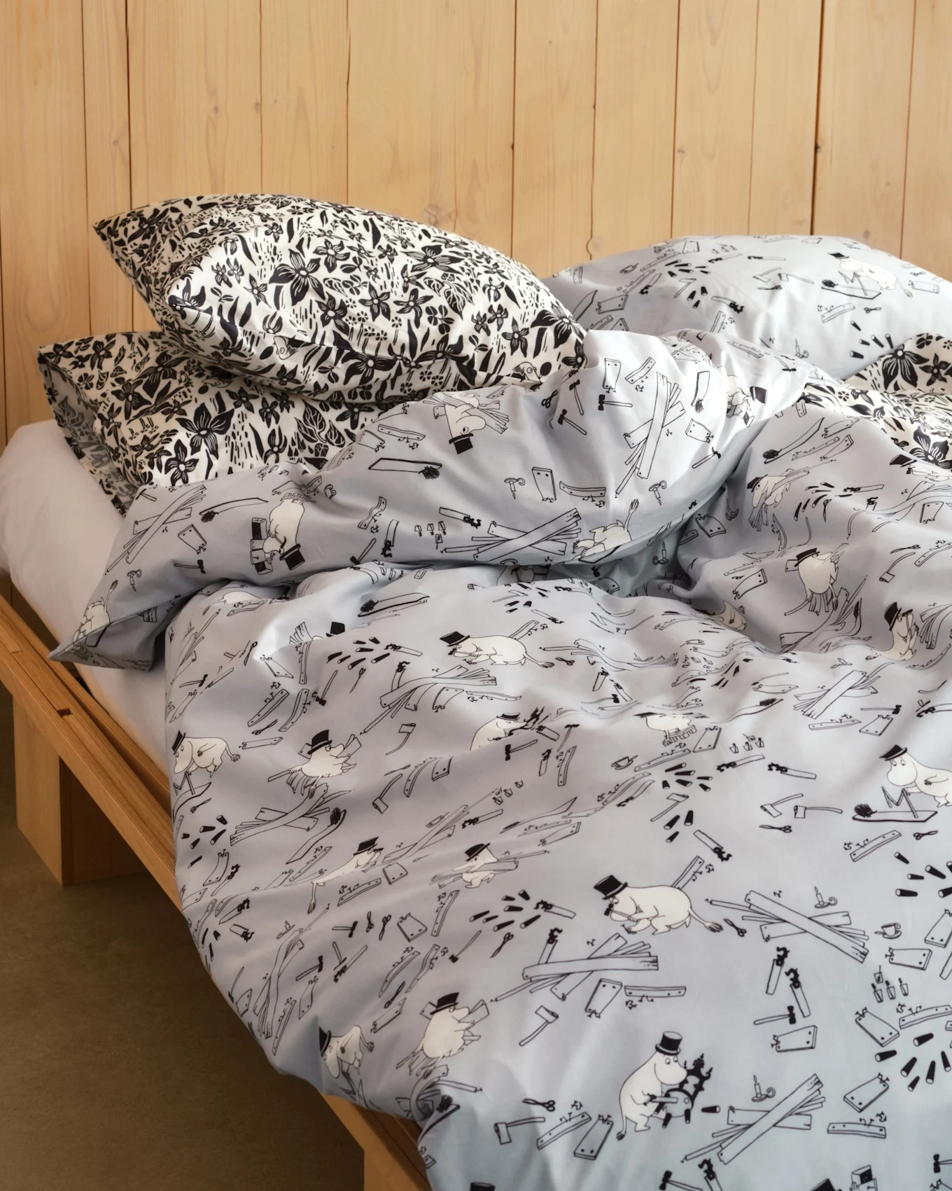 Cama con almohadas de estampado floral blanco y negro y edredón azul claro con figuras de dibujos animados, herramientas y tablas de madera. Somier de madera.