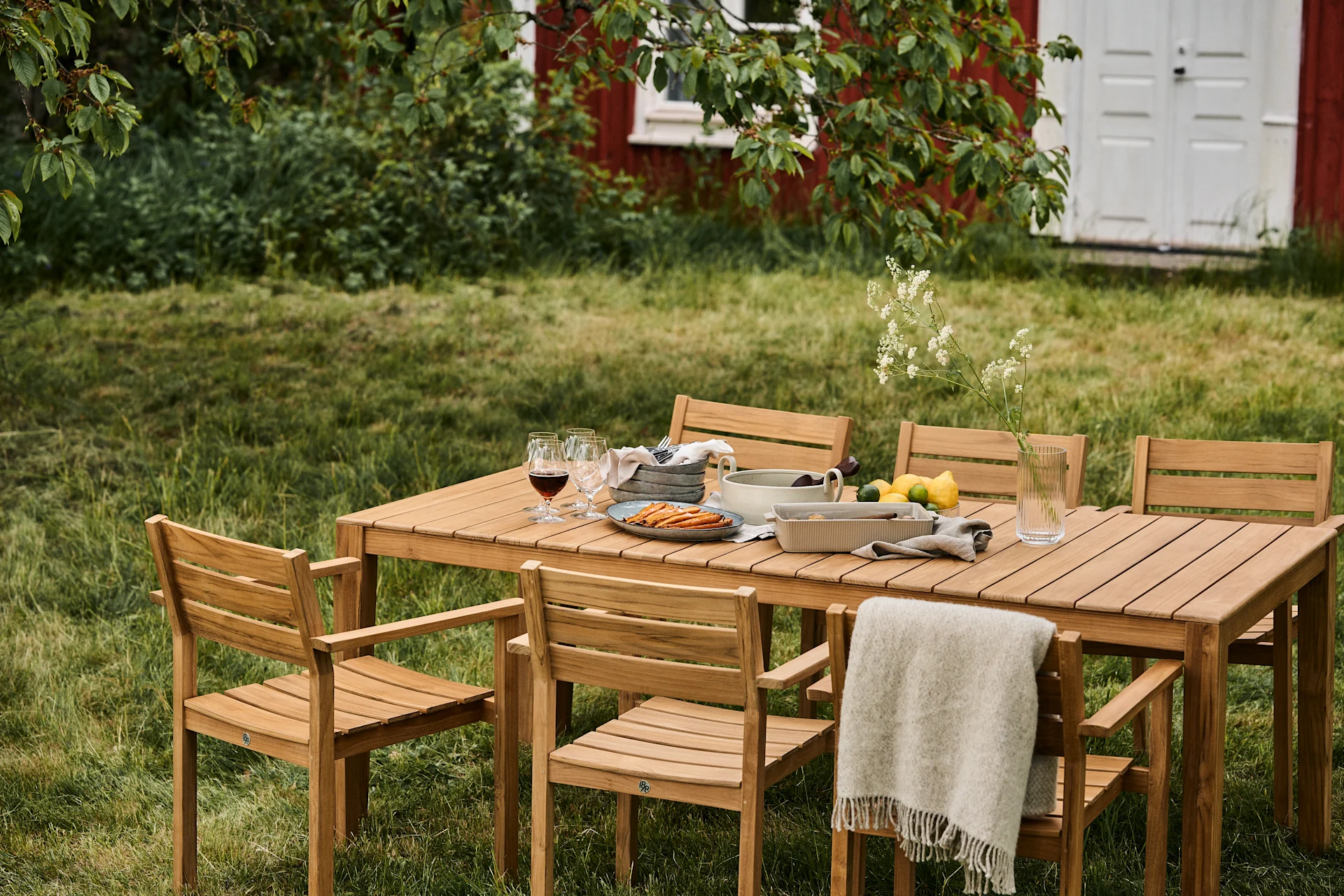 Conjunto de comedor exterior con mesa de madera y seis sillas en un césped, con comida, bebidas y una manta, con una casa roja al fondo.