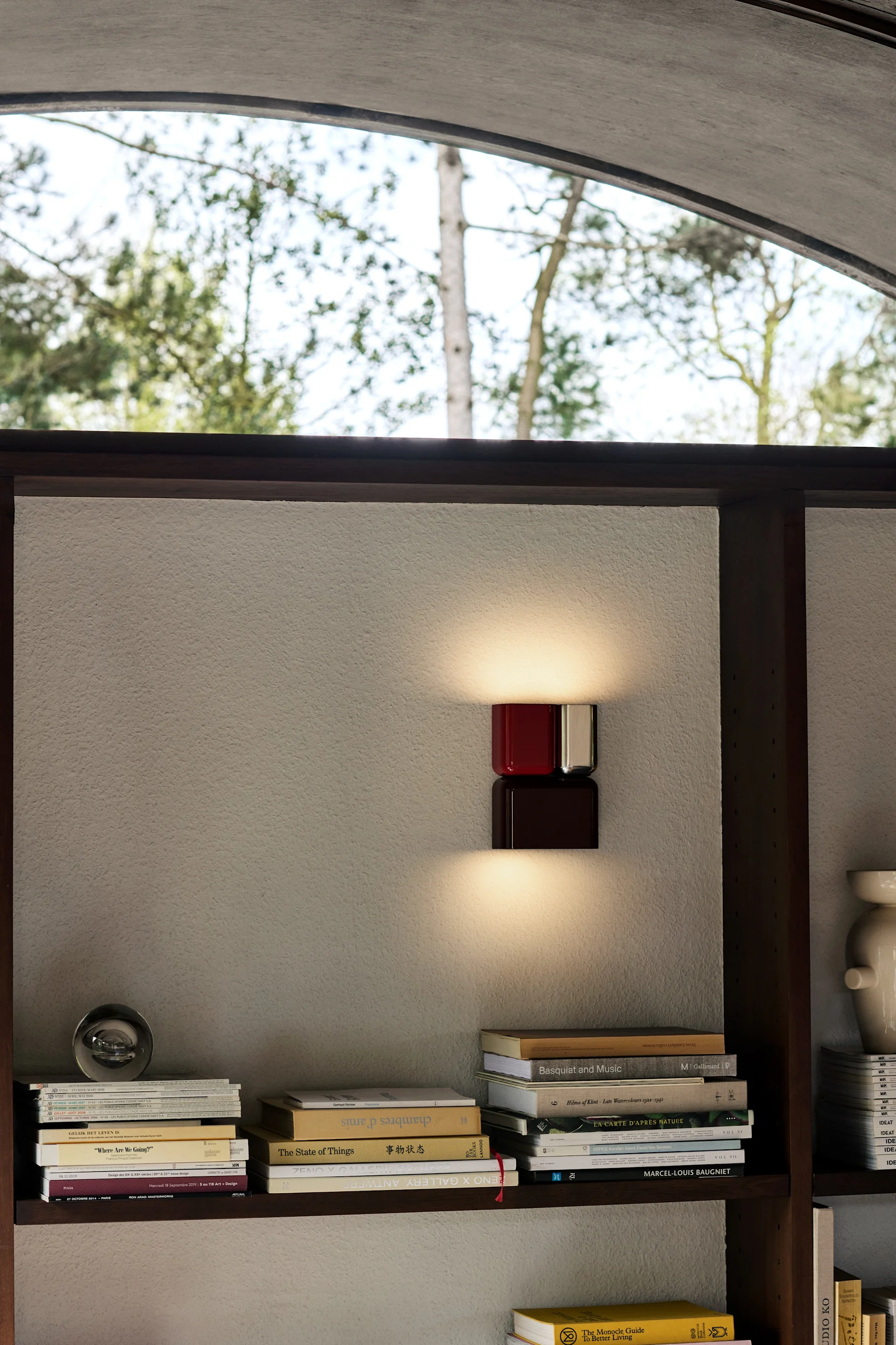 Una moderna lámpara de pared con un elemento superior rojo y otro inferior marrón oscuro en forma de cubo ilumina una pared texturizada entre estanterías repletas de libros. La lámpara de pared Tabata de &Tradition es perfecta para decorar paredes. 
