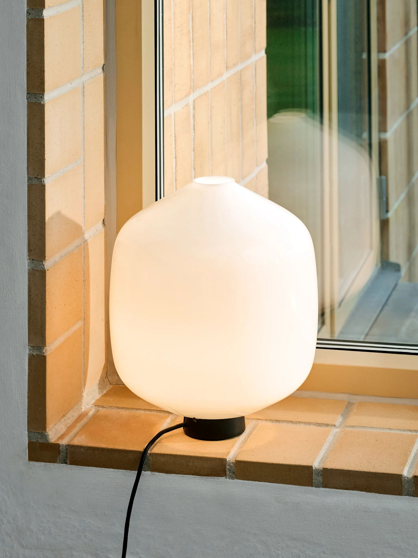 Una lámpara de mesa blanca, luminosa y redondeada con base y cable negros sobre un alféizar de ladrillo.