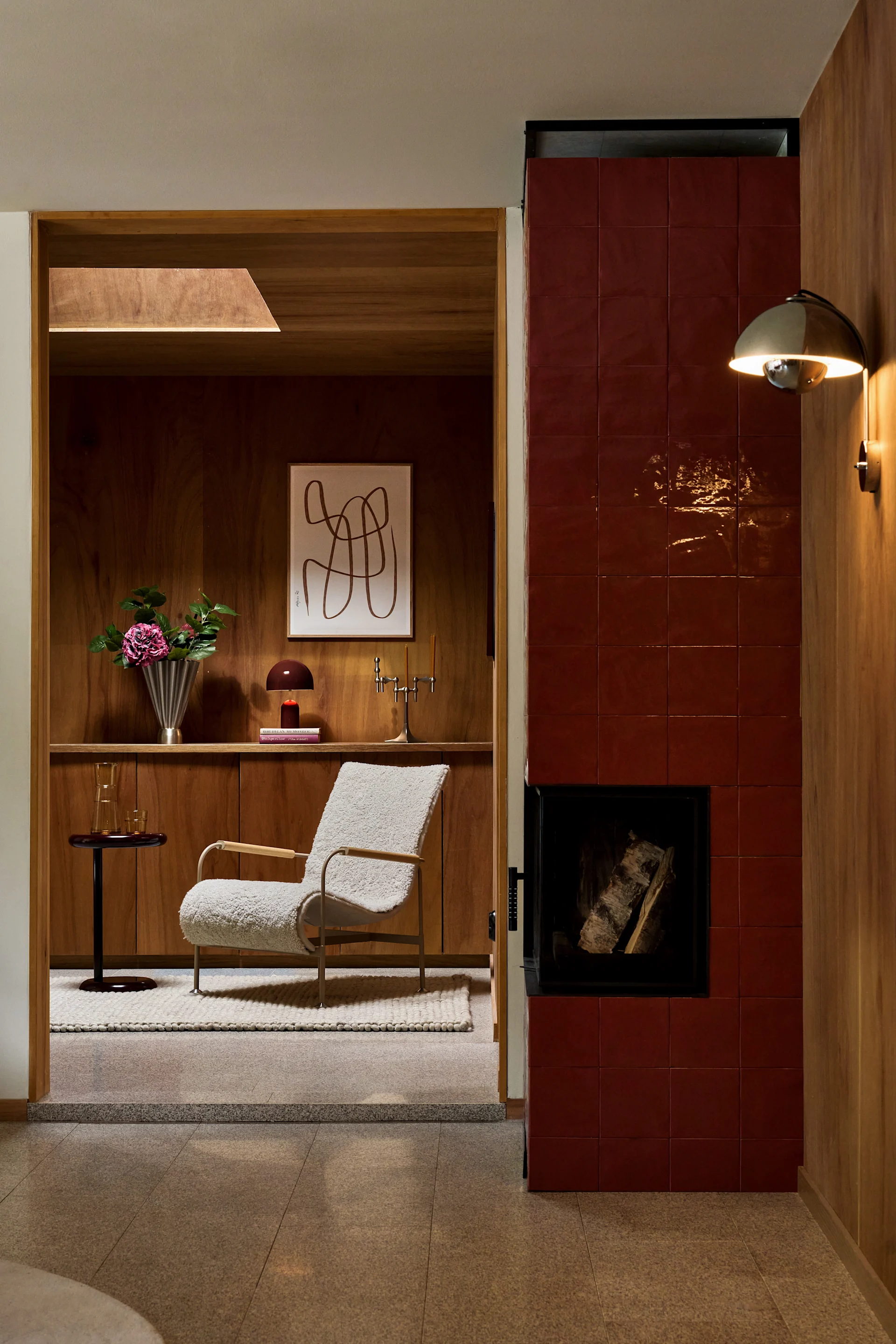 Una acogedora sala de estar con chimenea de azulejos rojos, un sillón bouclé crema y paneles de madera con estantes empotrados.