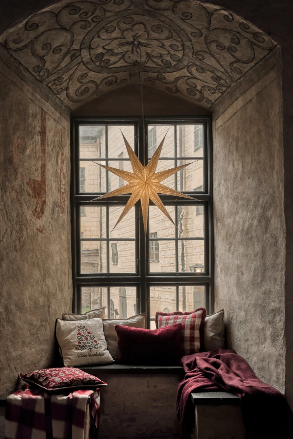 Un acogedor rincón con ventana en arco, con una estrella iluminada, cojines con motivos rojos y blancos y mantas sobre un asiento de piedra.