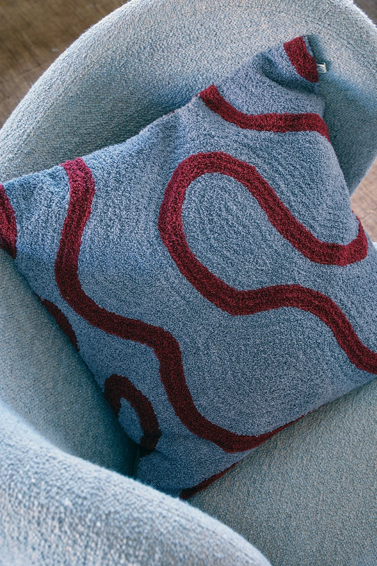 Un cojín azul claro texturizado con un patrón ondulado rojo oscuro descansando en un sillón azul claro.