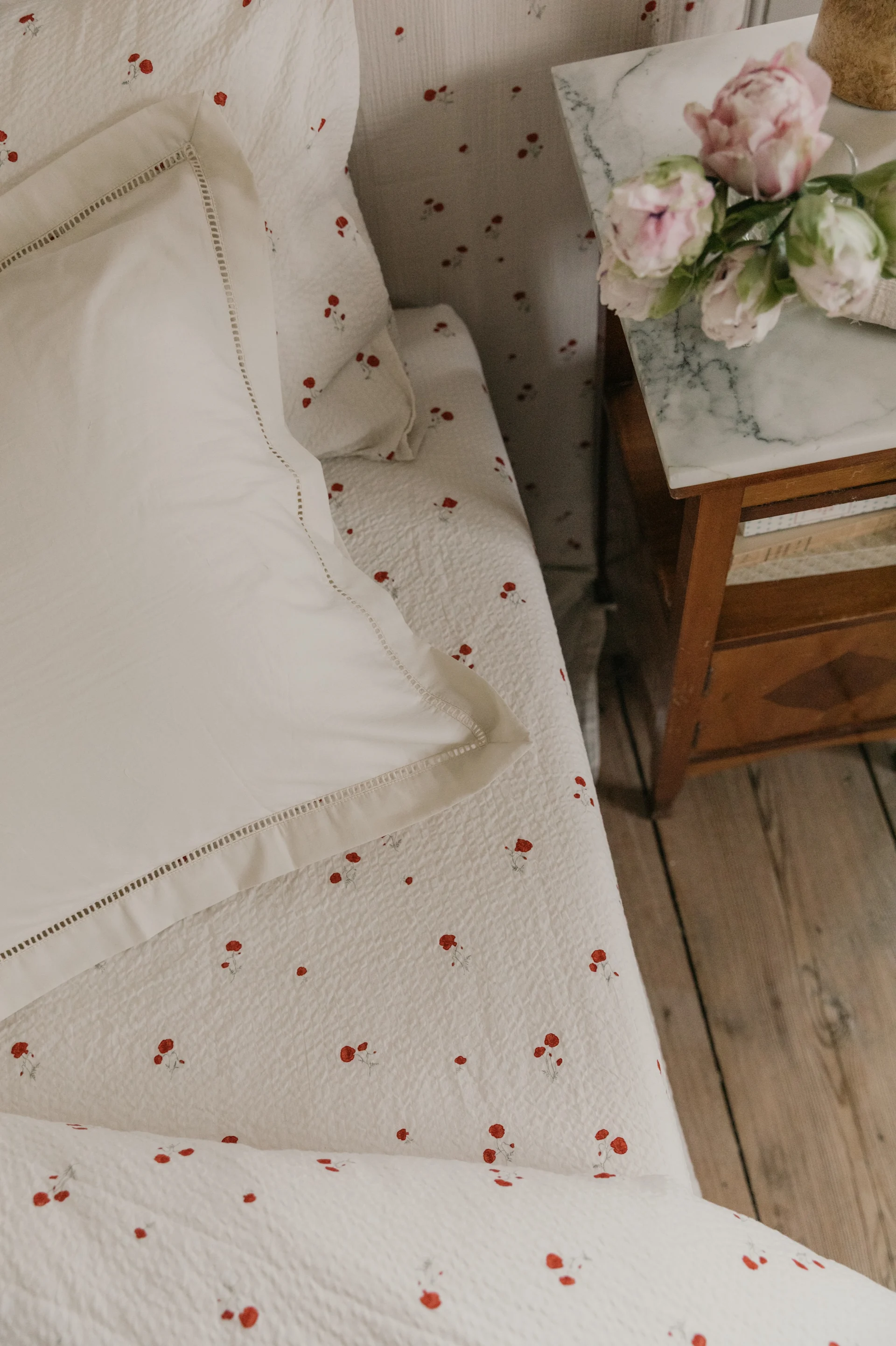 Ropa de cama blanca con estampado floral rojo, una almohada blanca y flores rosas en una mesita de noche de mármol.