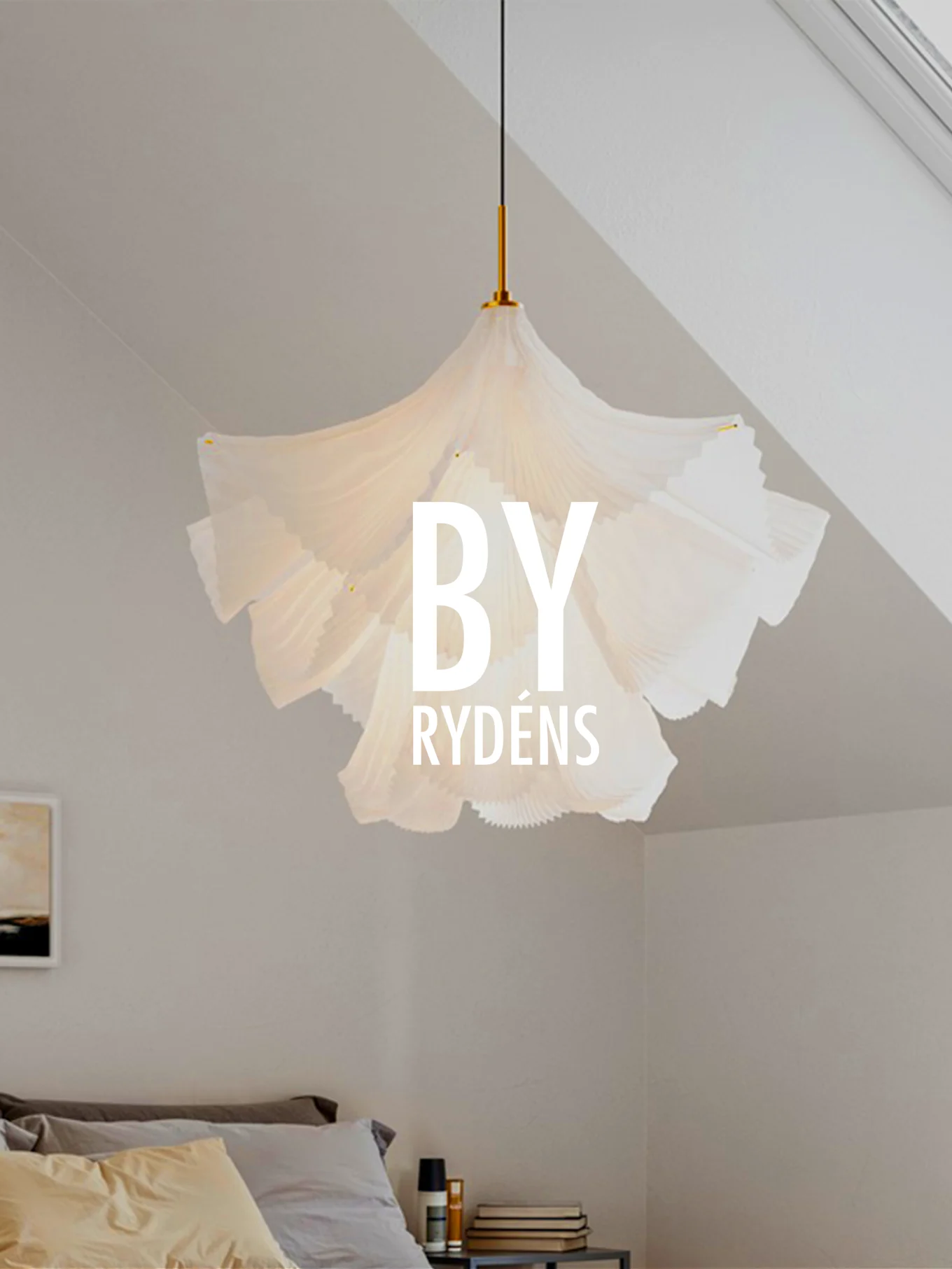 Una lámpara colgante escultural blanca y plisada cuelga sobre una cama con almohadas y una mesita de noche. 'BY RYDÉNS' está superpuesto en la lámpara.