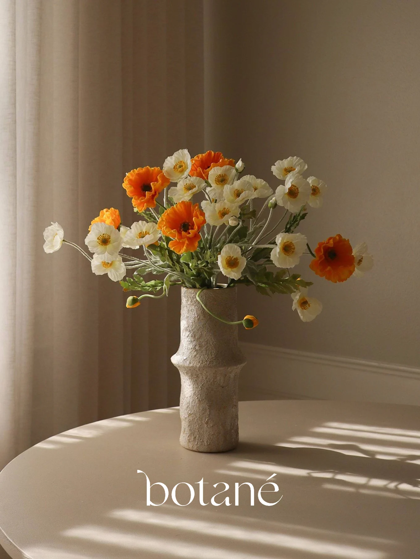 Un jarrón texturizado con un ramo de amapolas artificiales naranjas y blancas sobre una mesa con sombras rayadas.