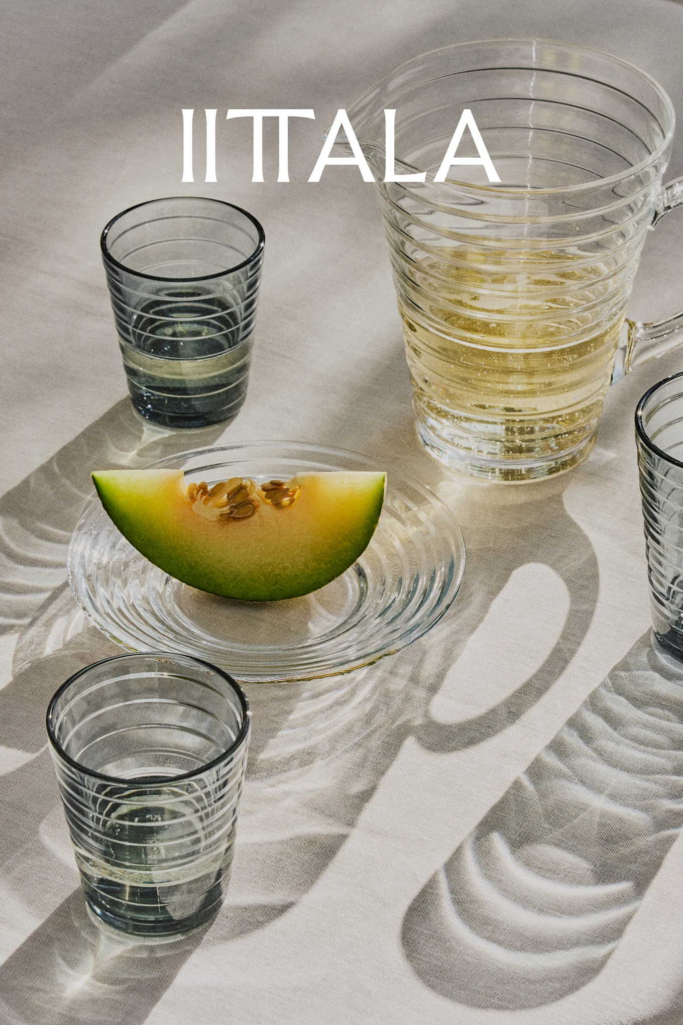 Logotipo IITTALA sobre vasos de cristal, una jarra con bebida espumosa y una rodaja de melón en un plato, todo sobre tela clara.