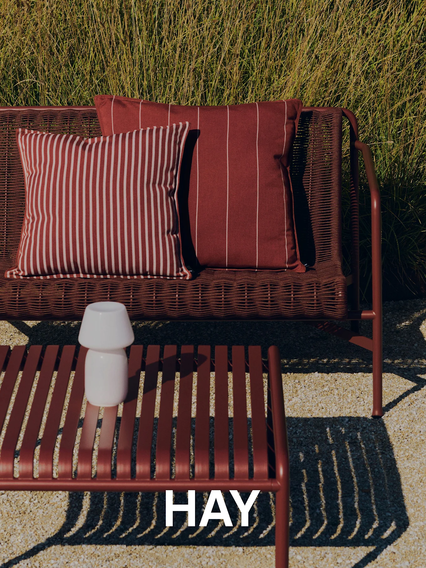 Escena al aire libre con un sofá rojo trenzado, cojines a rayas, una mesa de centro de lamas con una lámpara blanca y hierba alta al fondo. Logo de HAY.