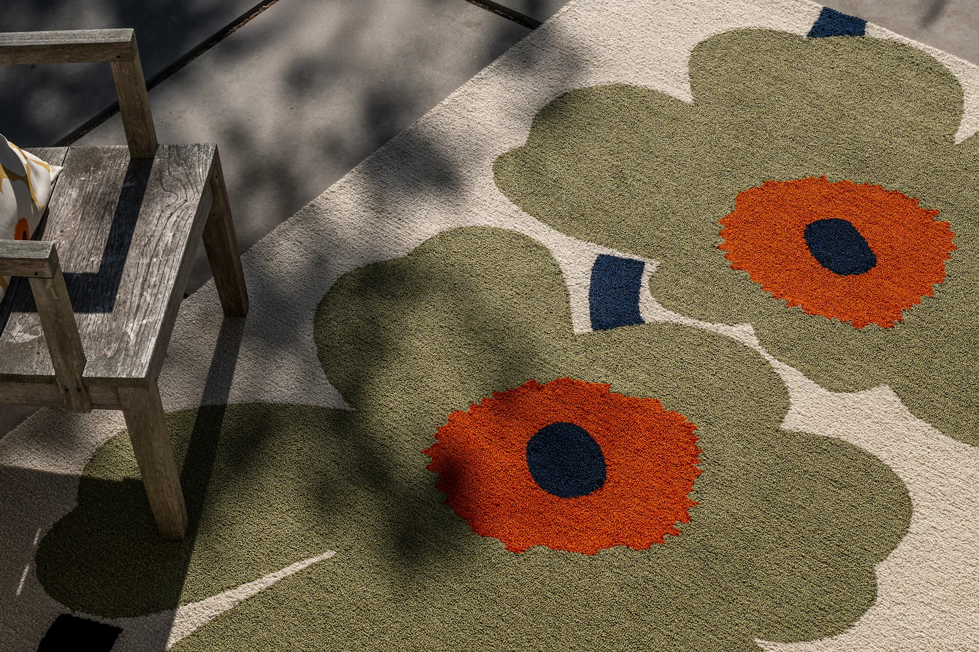 Alfombra exterior con patrón floral grande en verde oliva, naranja y azul oscuro, junto a una silla de madera rústica bajo luz solar moteada.