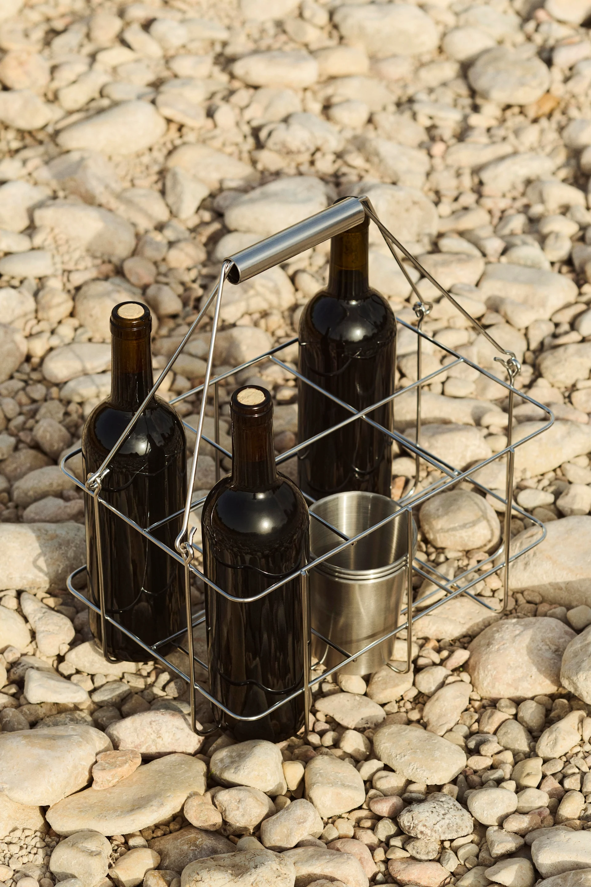 Tres botellas de vino y una copa de metal en un portabotellas sobre un lecho de guijarros.
