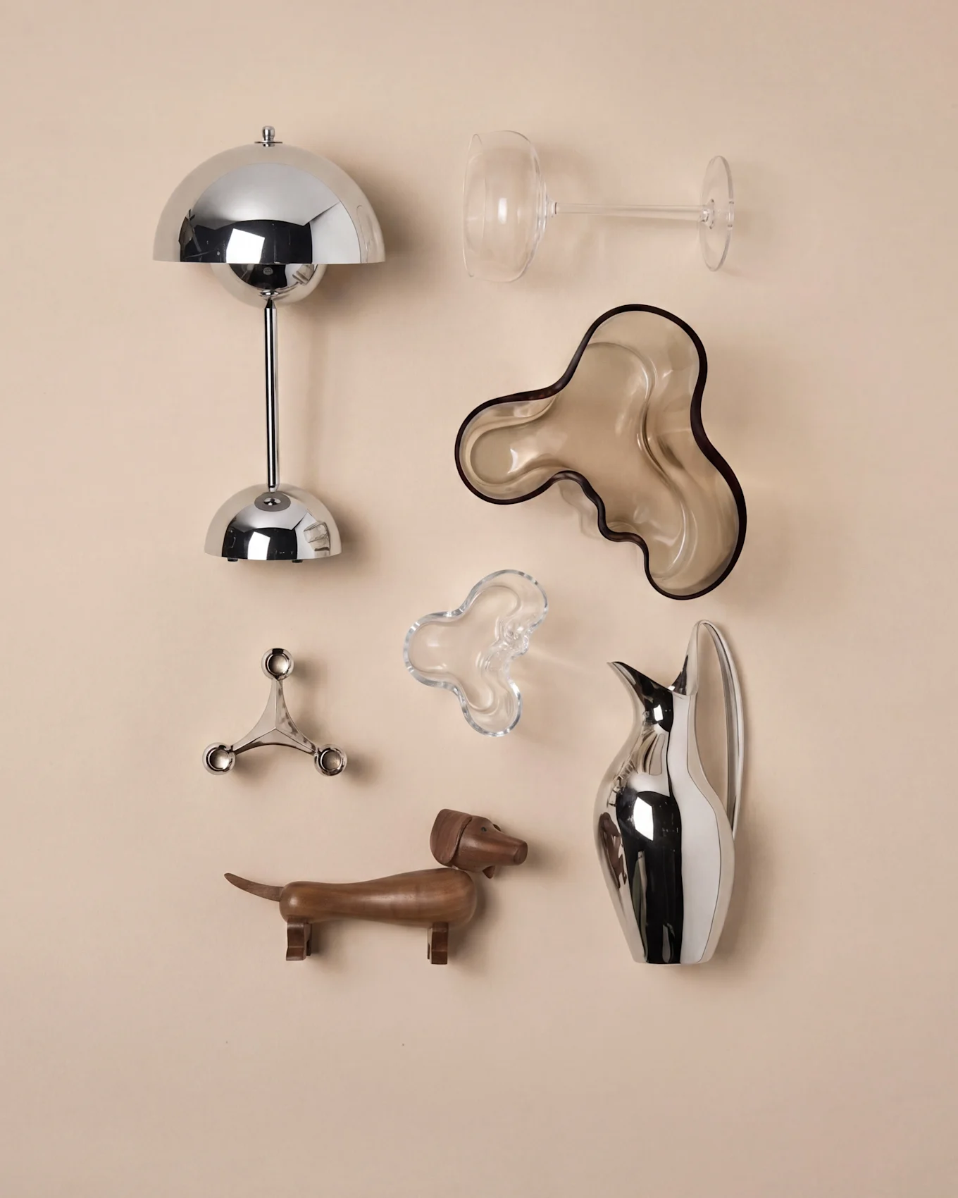 Un conjunto de objetos decorativos: una lámpara cromada, copa de vino, dos cuencos orgánicos, un candelabro, un perro salchicha de madera y una jarra.