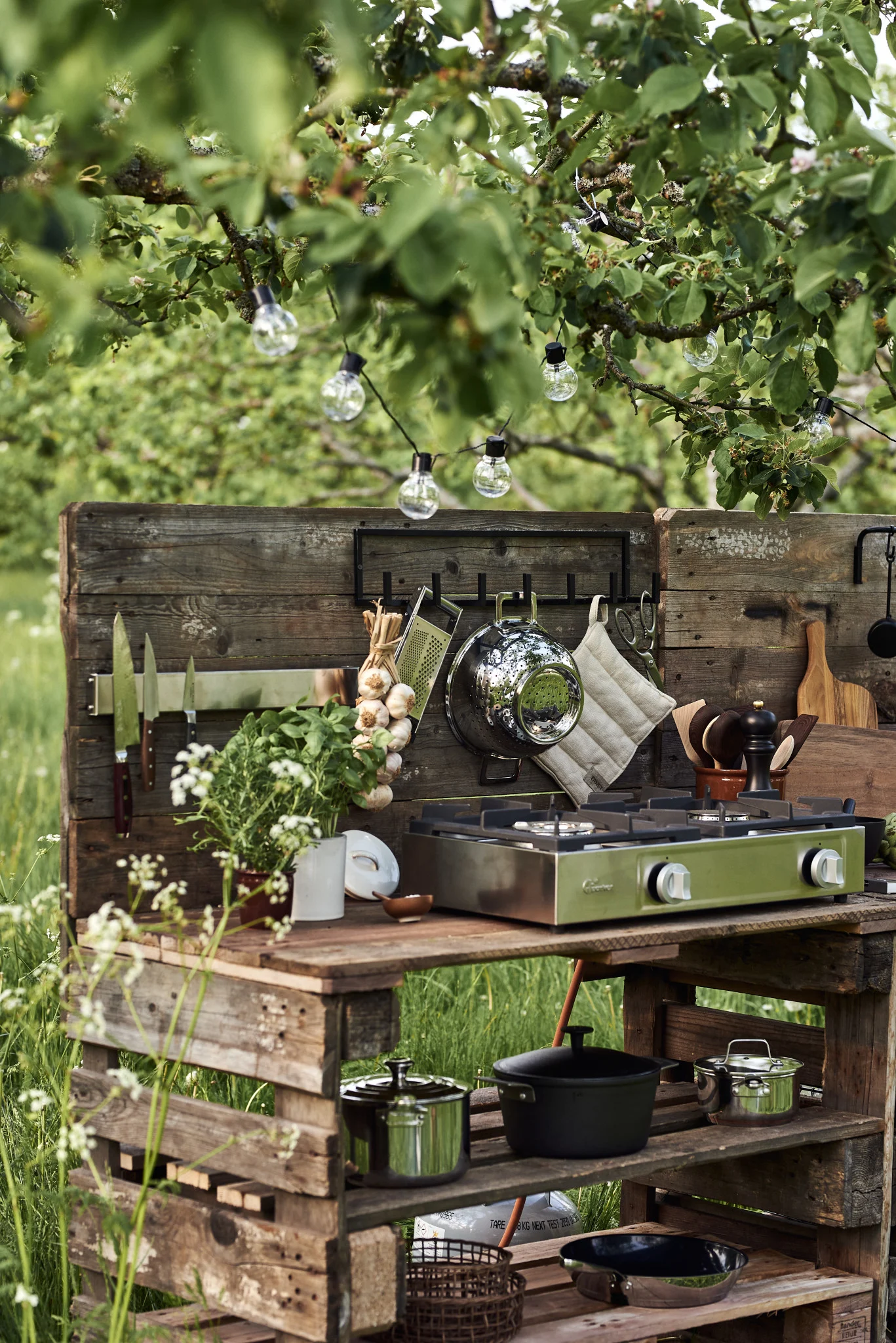 Una cocina exterior hecha de paletas de madera, con una estufa de gas verde de dos quemadores, utensilios de cocina, hierbas y luces de cadena.