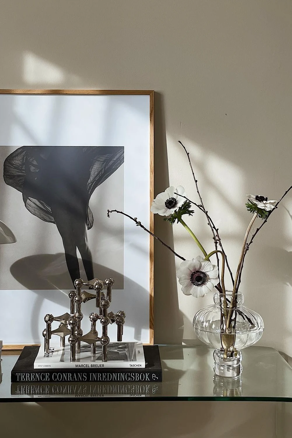 Foto enmarcada en blanco y negro de piernas con tela fluida, candelabro modular plateado, libros y anémonas en jarrón.