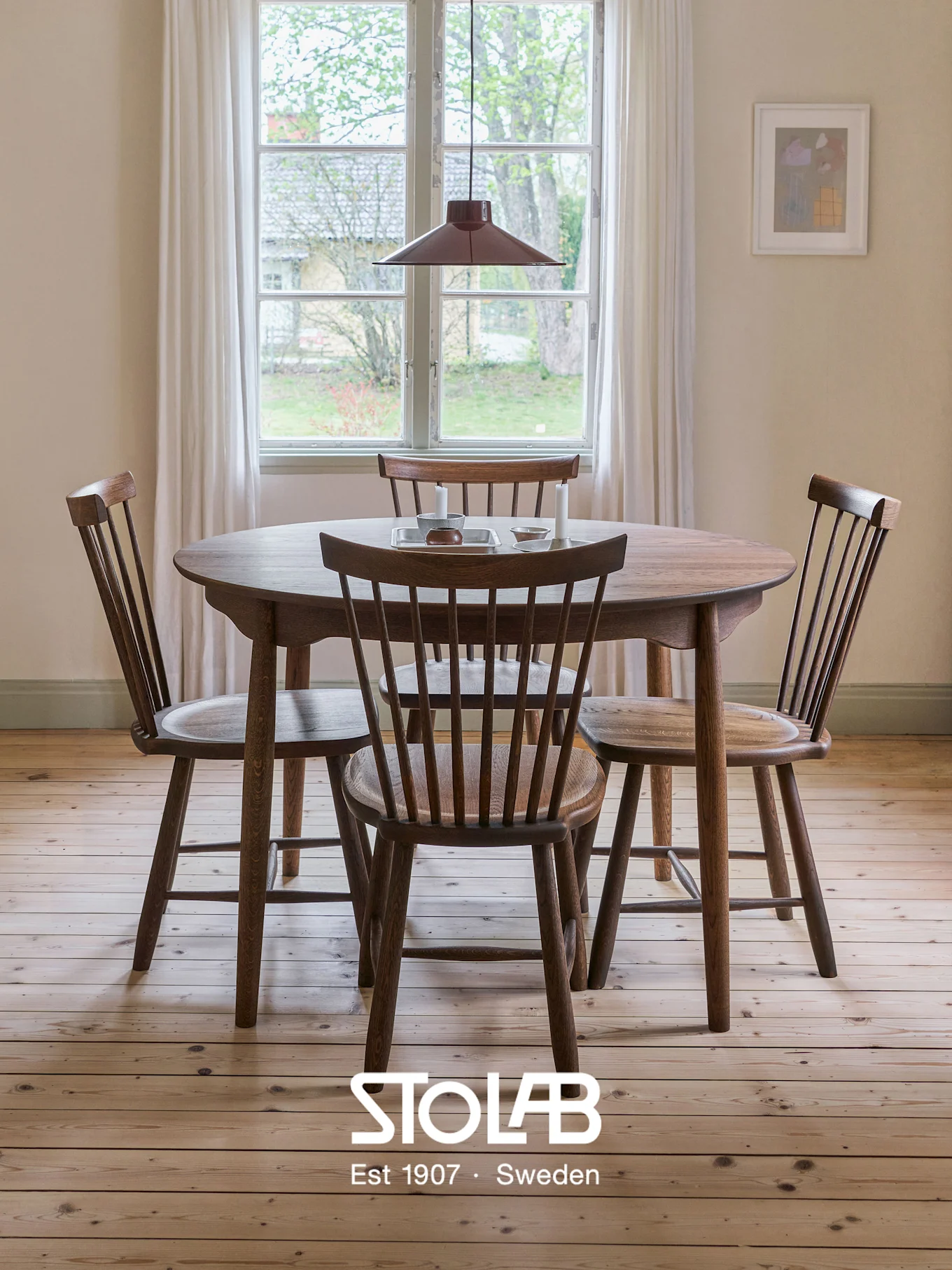 Mesa de comedor redonda de madera con cuatro sillas de respaldo de huso en una habitación luminosa, suelo de madera clara, ventana y lámpara colgante roja.