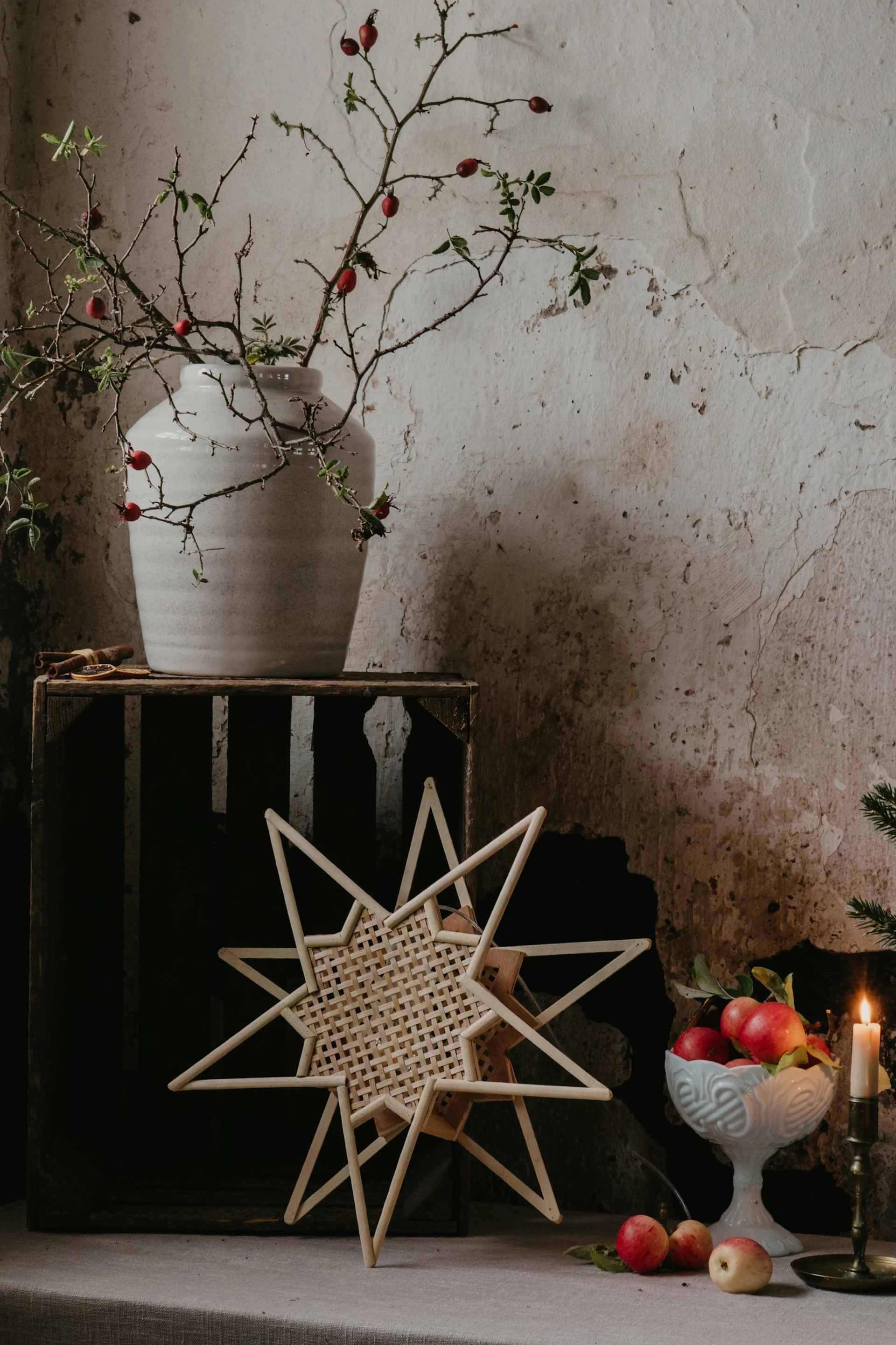 Una lámpara con forma de estrella cuelga frente a una ventana, sobre un asiento acolchado con velas, una taza y pasteles.