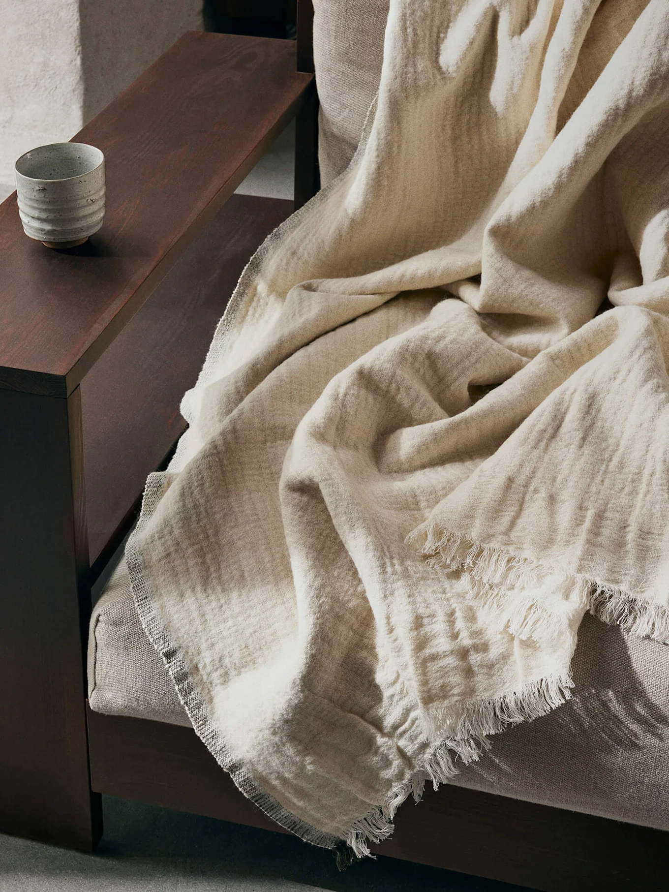 Manta texturizada beige claro con bordes deshilachados sobre un sofá, con una taza de cerámica en un reposabrazos de madera oscura.