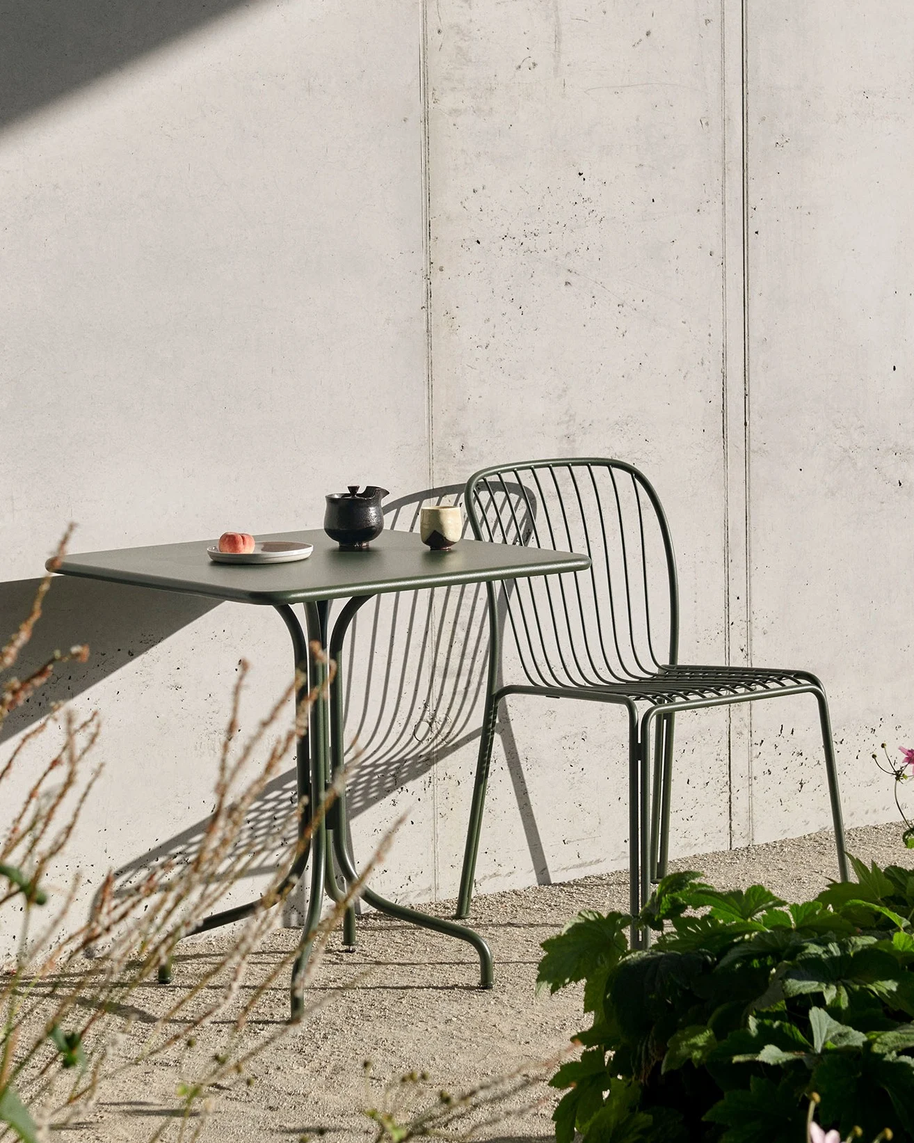 Una mesa exterior verde oscuro con un melocotón, tetera y taza, y una silla de listones a juego, contra una pared de hormigón con sombras marcadas.