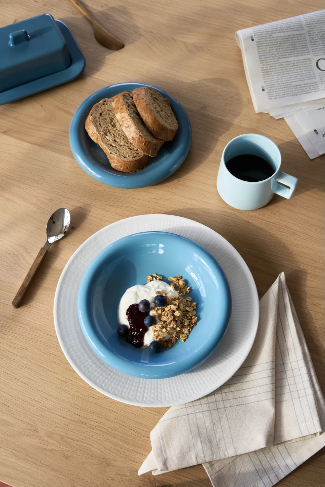 Desayuno con yogur, granola, arándanos y pan en platos azules y blancos, café en taza azul, mantequillera en mesa de madera.