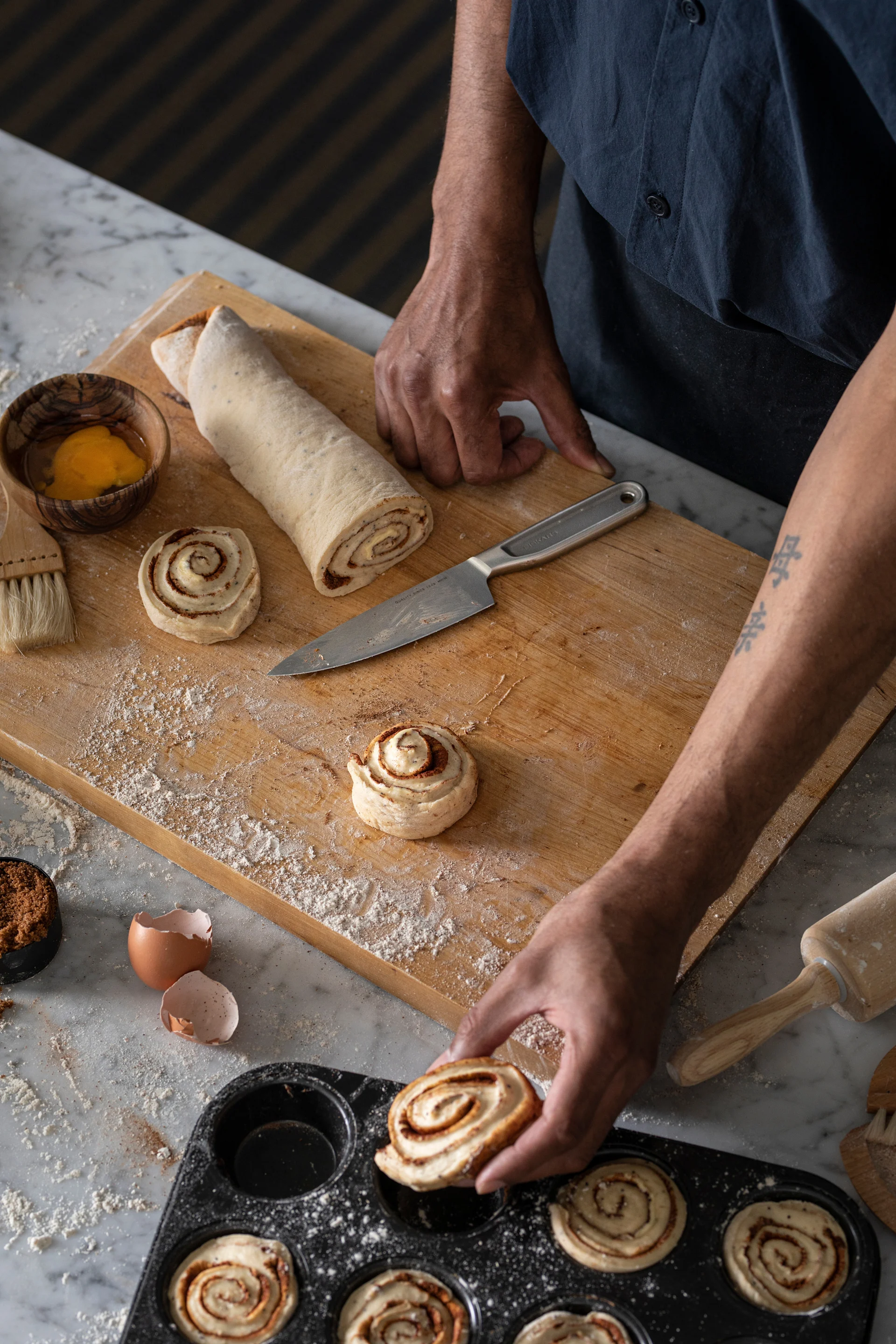 Manos colocando un rollo de canela en un molde para muffins, con masa, harina e ingredientes en una encimera de mármol.