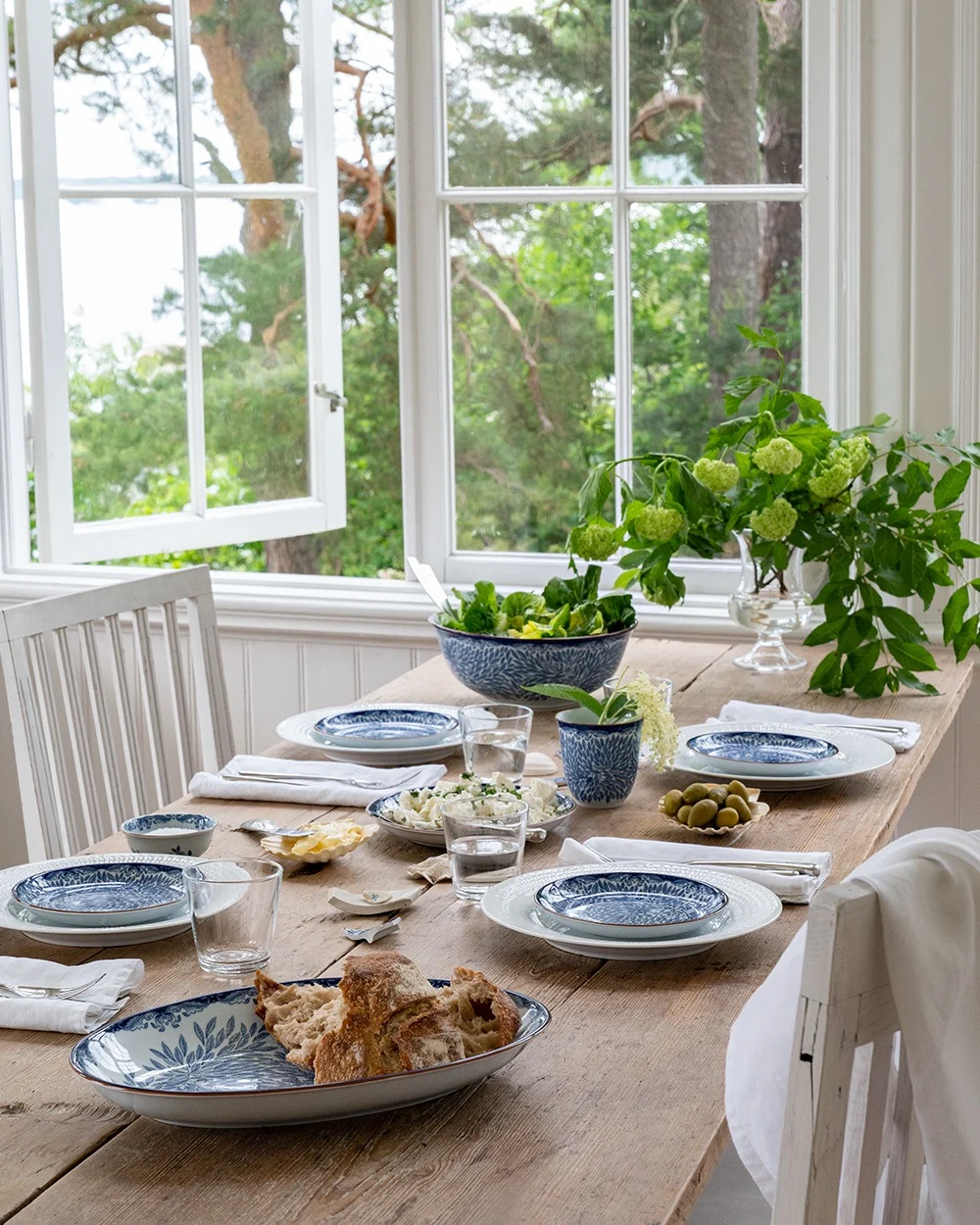 Una mesa de comedor rústica de madera con vajilla azul y blanca, ensalada, pan y aceitunas, junto a una ventana abierta con vistas a árboles verdes y agua.