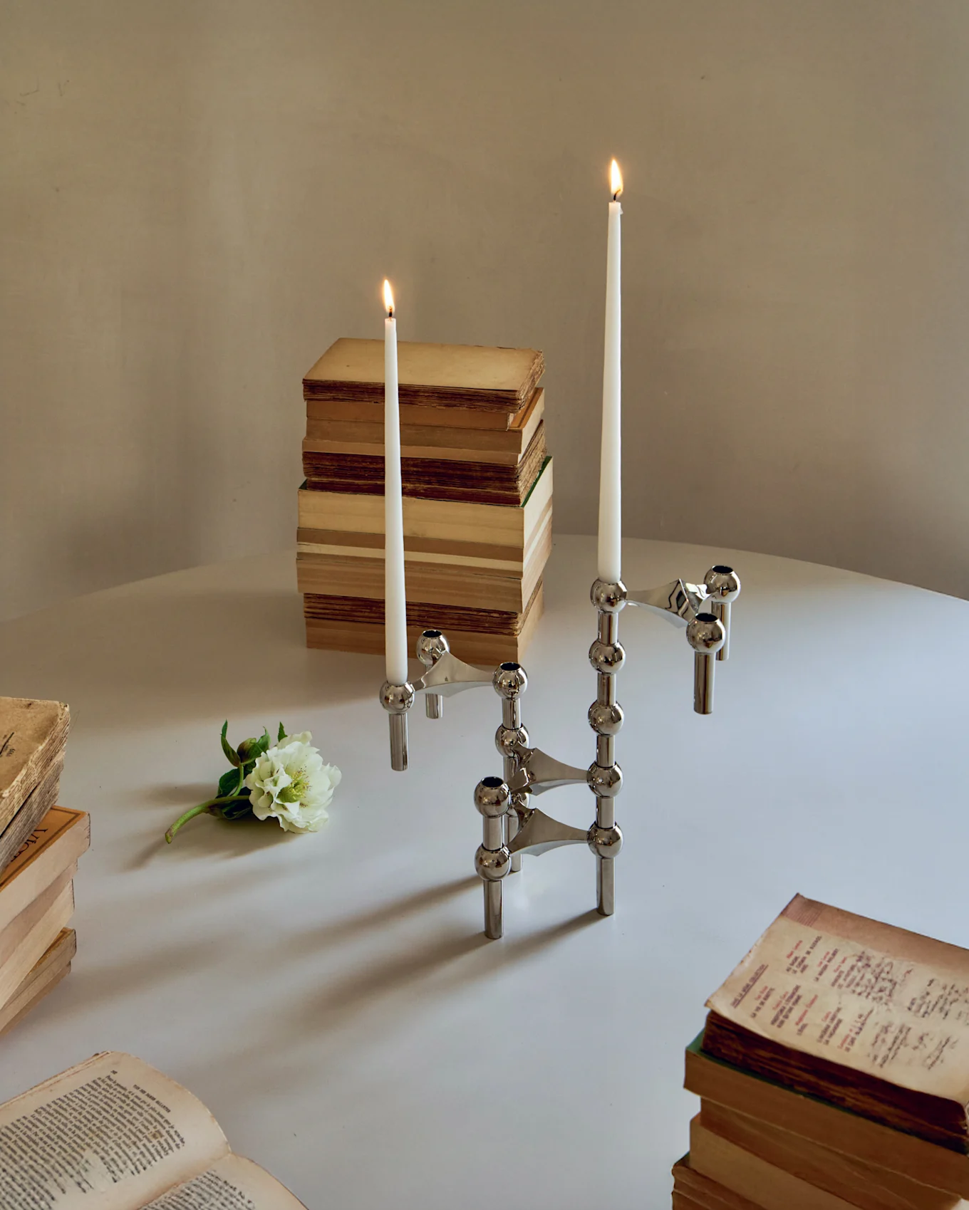 Dos candelabros modulares cromados con velas blancas encendidas, rodeados de libros antiguos y una flor blanca sobre una mesa.