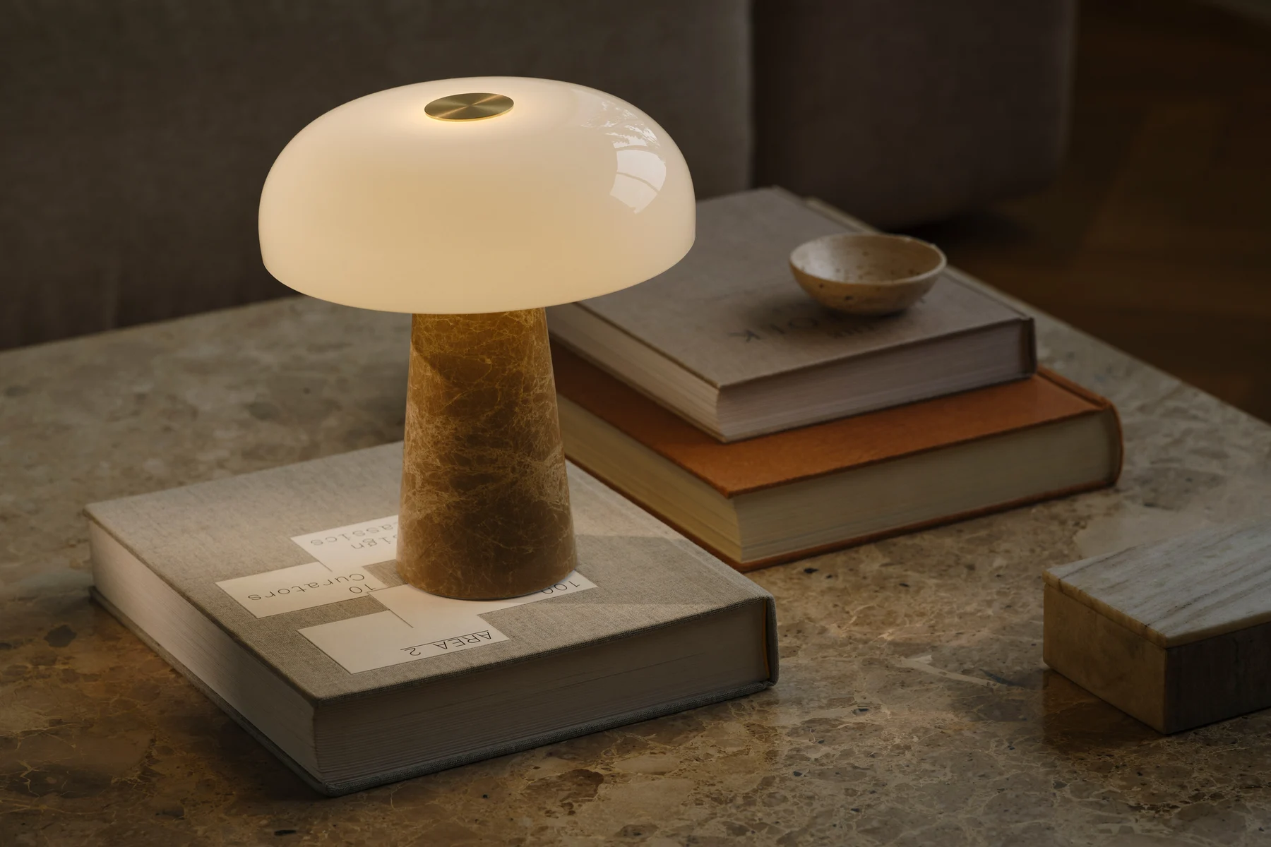Lámpara de mesa en forma de hongo con pantalla de cristal blanco y base cónica de mármol sobre una mesa de mármol con libros apilados.