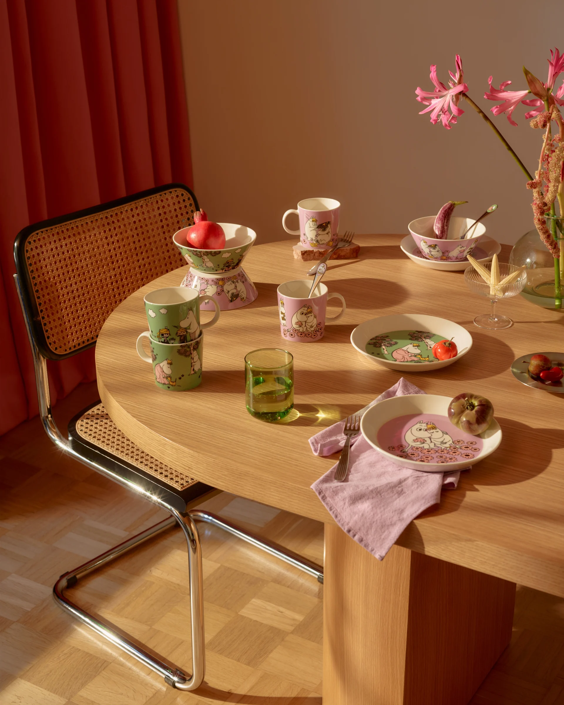 Mesa de comedor con vajilla de Moomin rosa y verde, frutas frescas, un vaso verde y flores rosas.