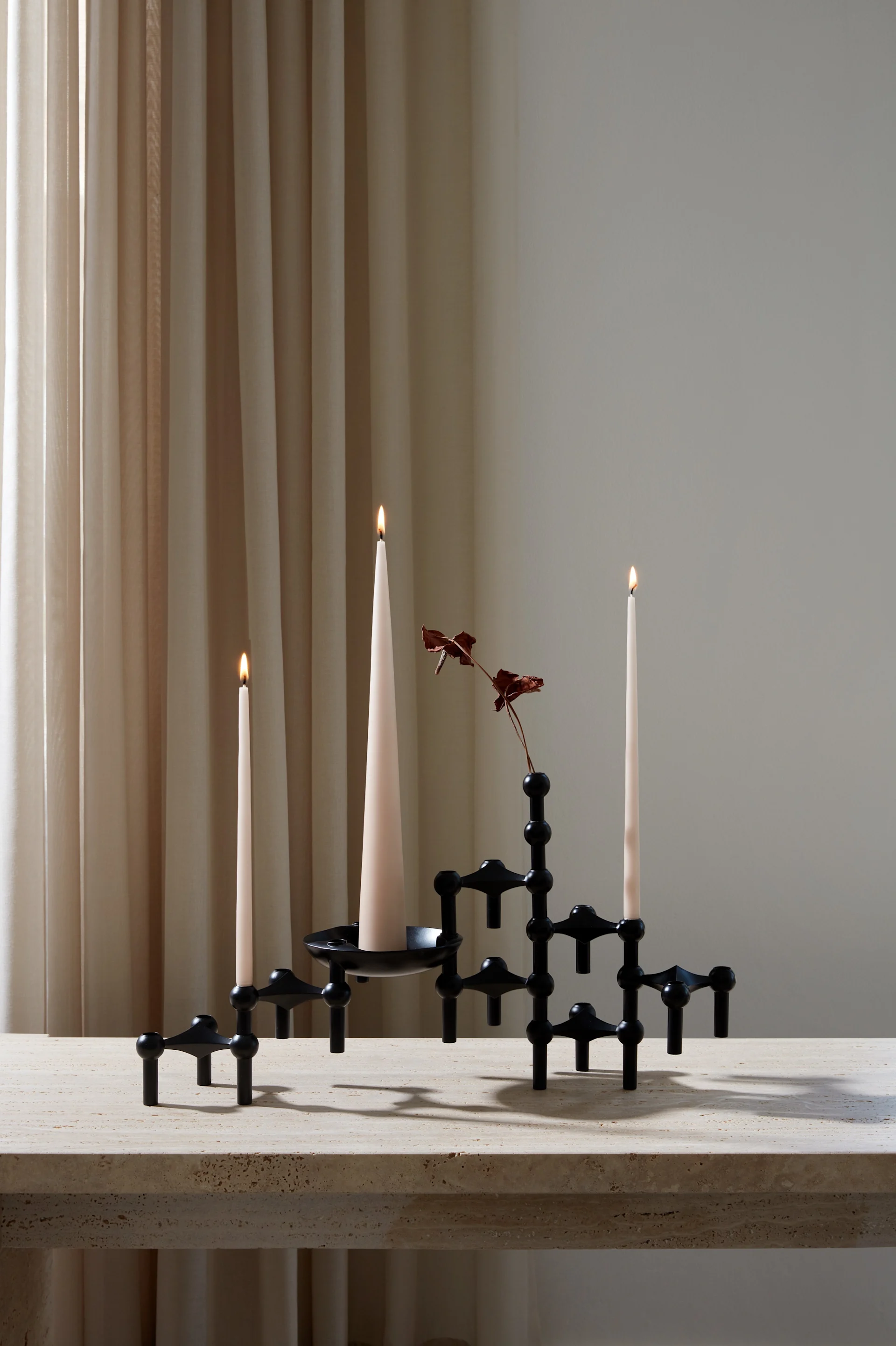 Candelabro modular negro con tres velas cónicas encendidas de color crema y una rama seca roja sobre una mesa de piedra beige, con cortinas claras.