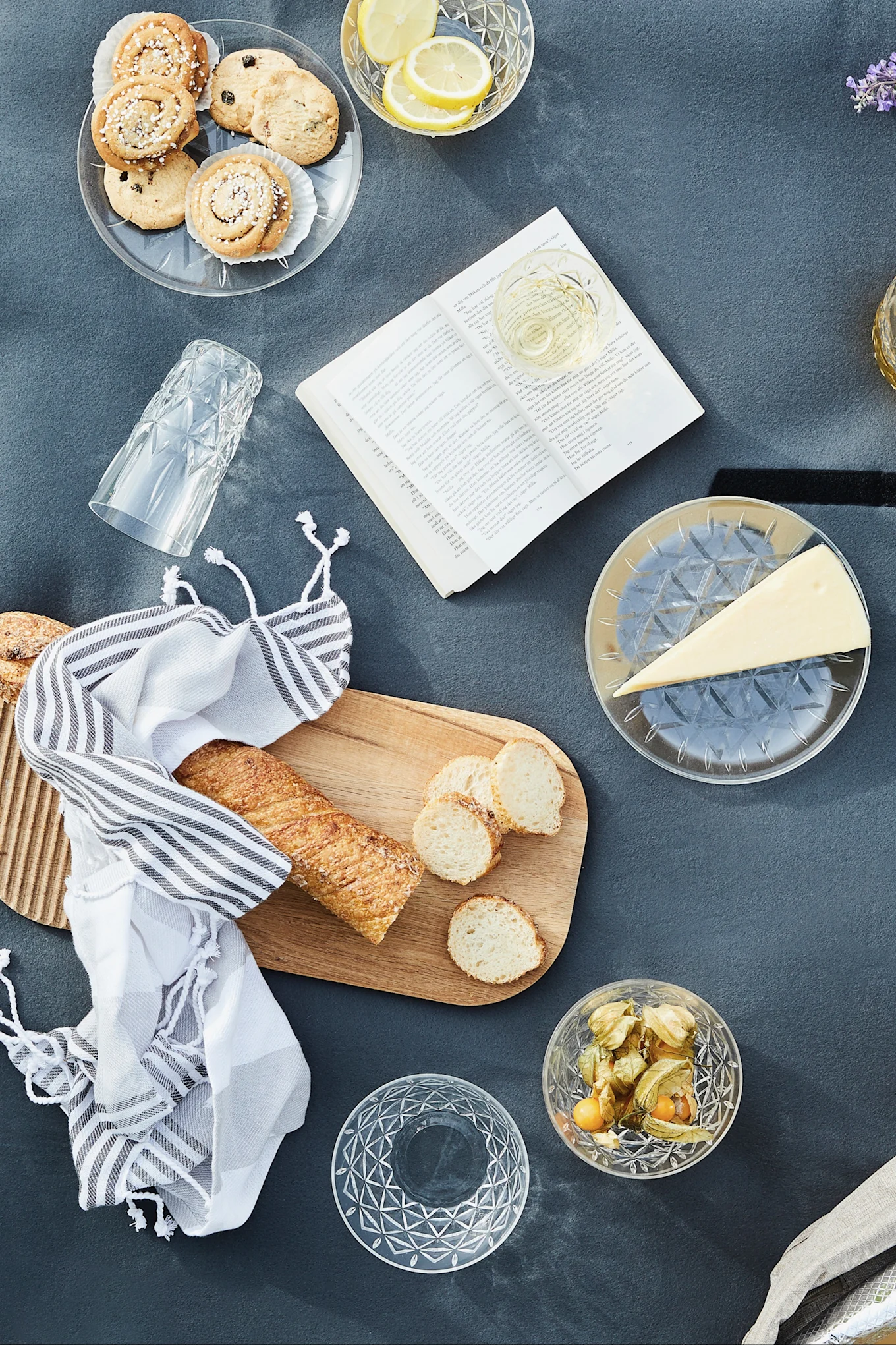 Vista aérea de un picnic con galletas, limones, vino, queso, baguette, physalis y un libro abierto sobre una manta gris.