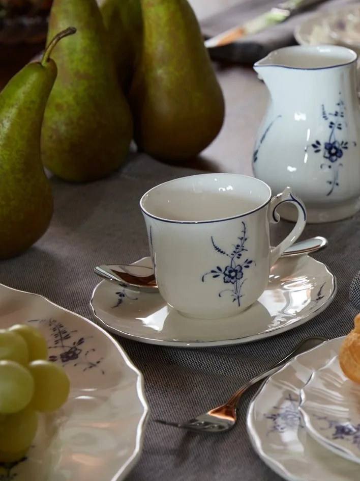 Taza, platillo y jarra de porcelana blanca con estampado floral azul en una mesa con peras y uvas.